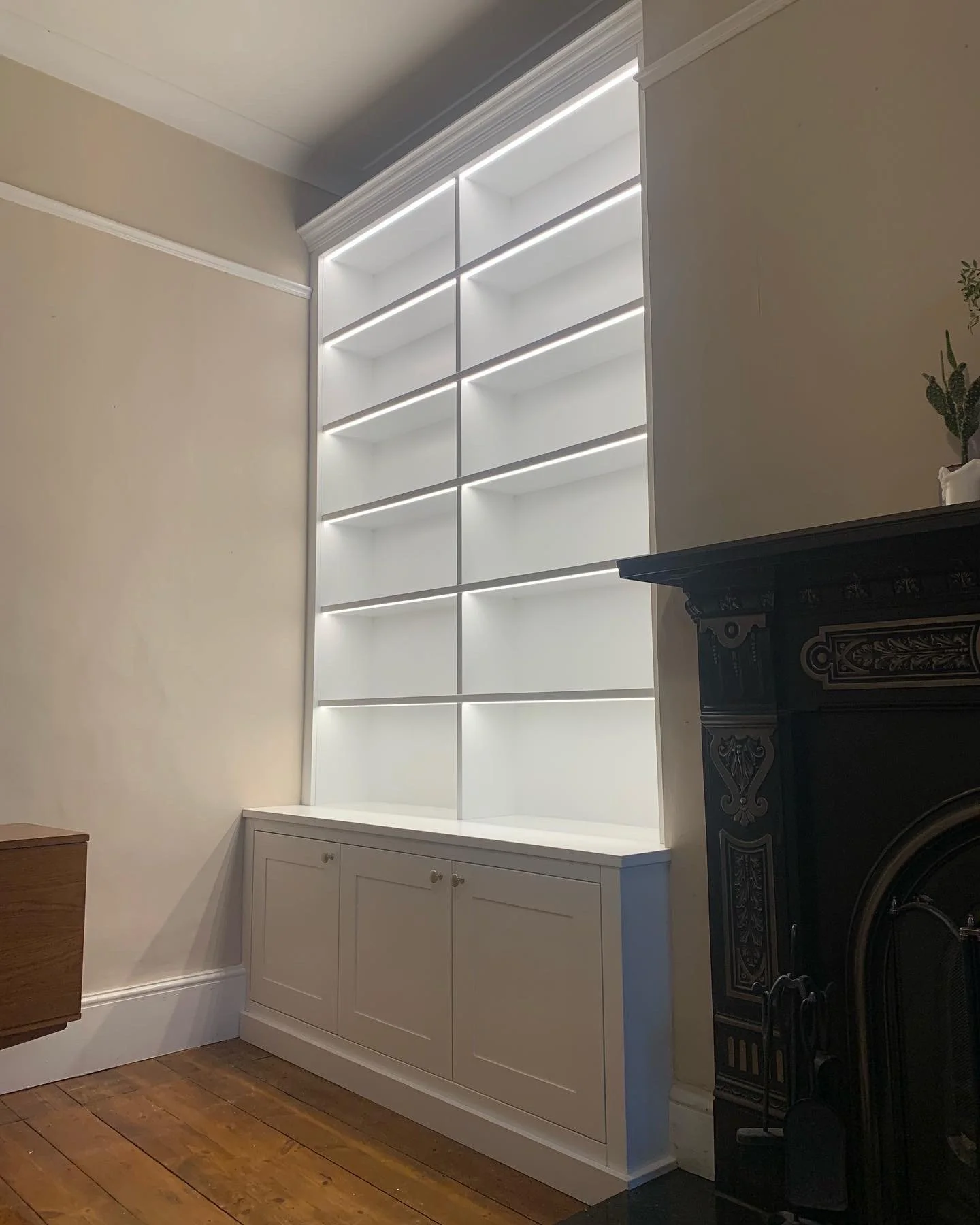 White built-in bookshelf with LED lighting, white cabinetry, against a beige wall, next to a black fireplace with decorative detailing, on a wooden floor.