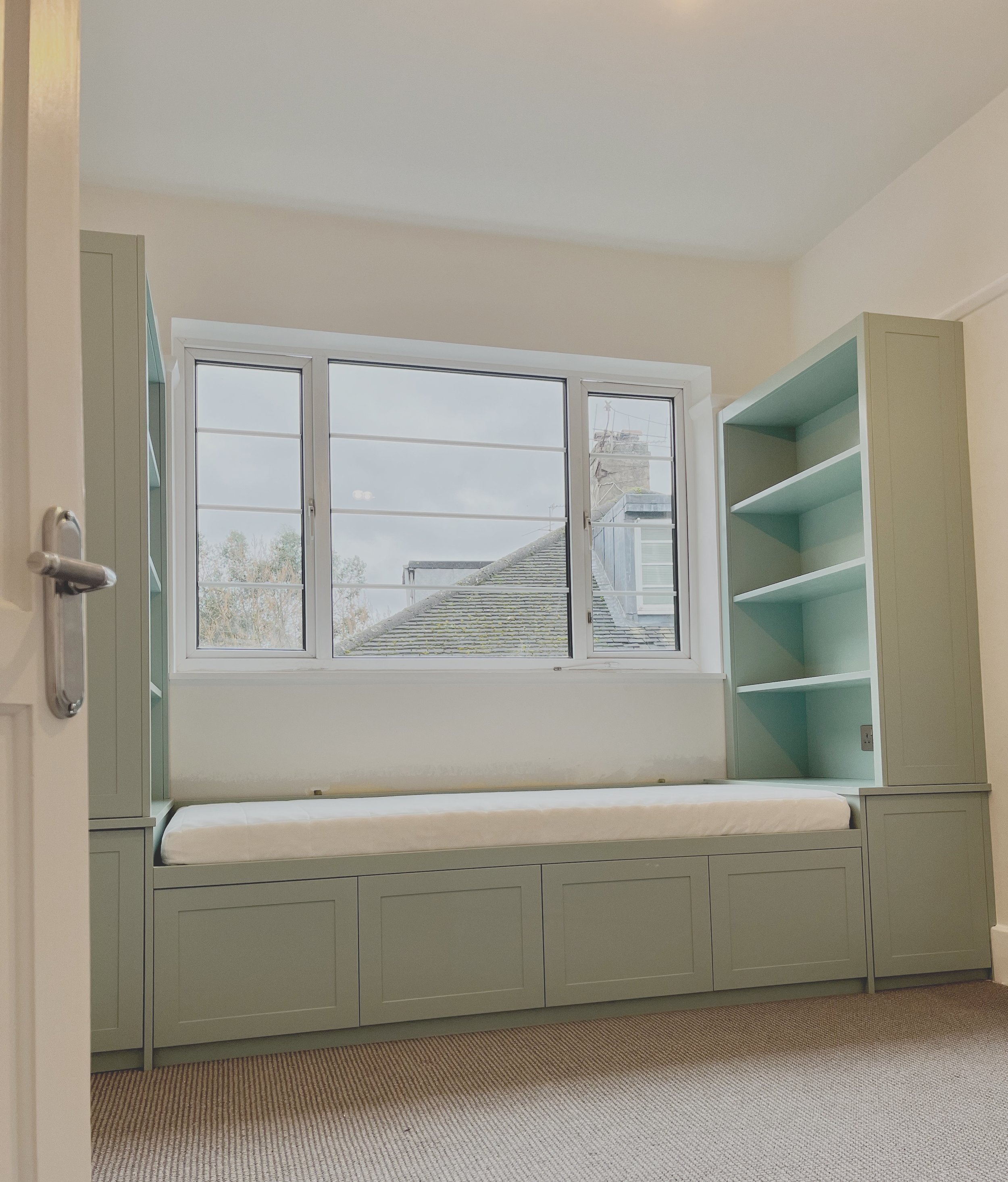Light green built-in window seat with storage underneath, positioned beneath a large window, in a room with beige carpet and white walls.