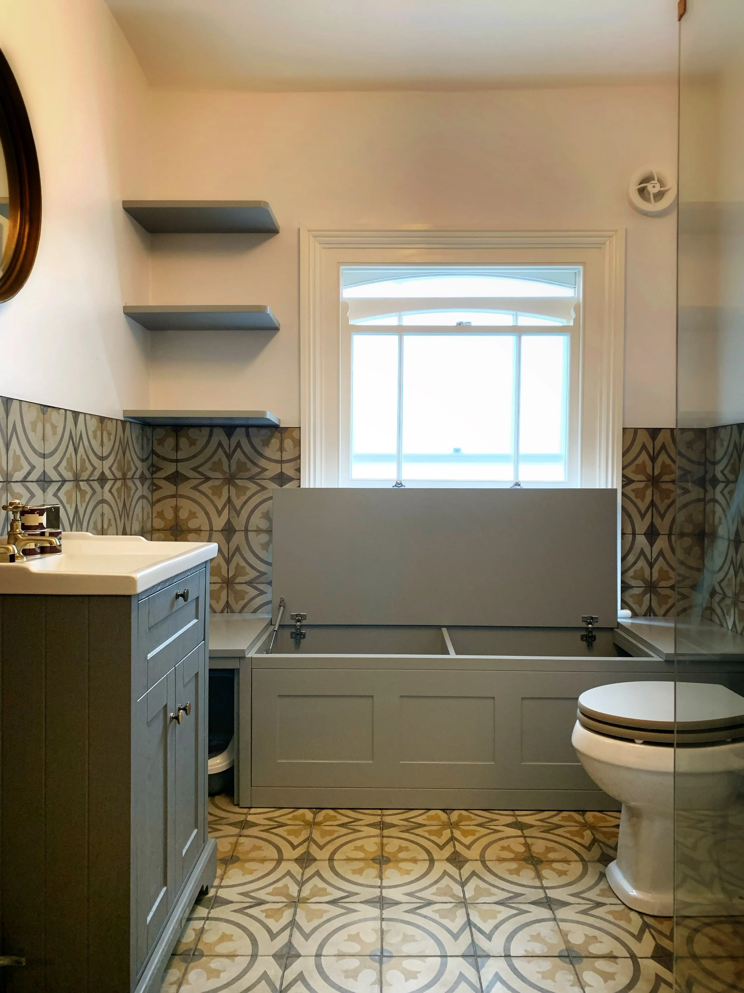 Bathroom with patterned floor tiles, a vanity with a sink and gold fixtures, wall-mounted floating shelves, a bathtub with a fold-down cover, a window with blinds, and a toilet.