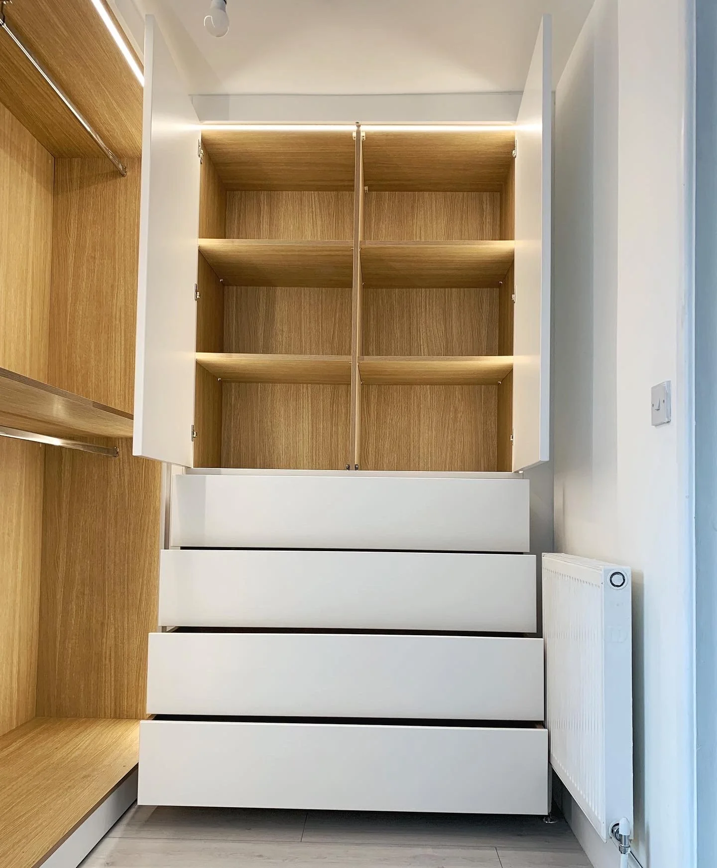 Open wooden closet with four empty shelves, white drawers below, and white doors on each side. A radiator is on the right side, and a light switch is visible on the wall.