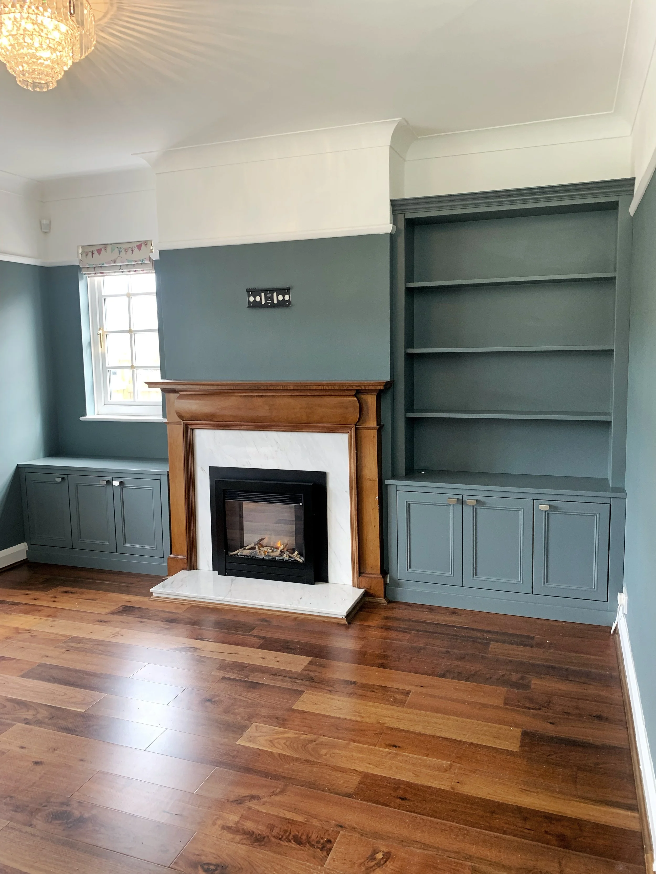 Living room with teal walls, a fireplace with wooden mantel and marble surround, built-in bookshelves and cabinets, a window with a decorative valance, hardwood floors, and a chandelier.