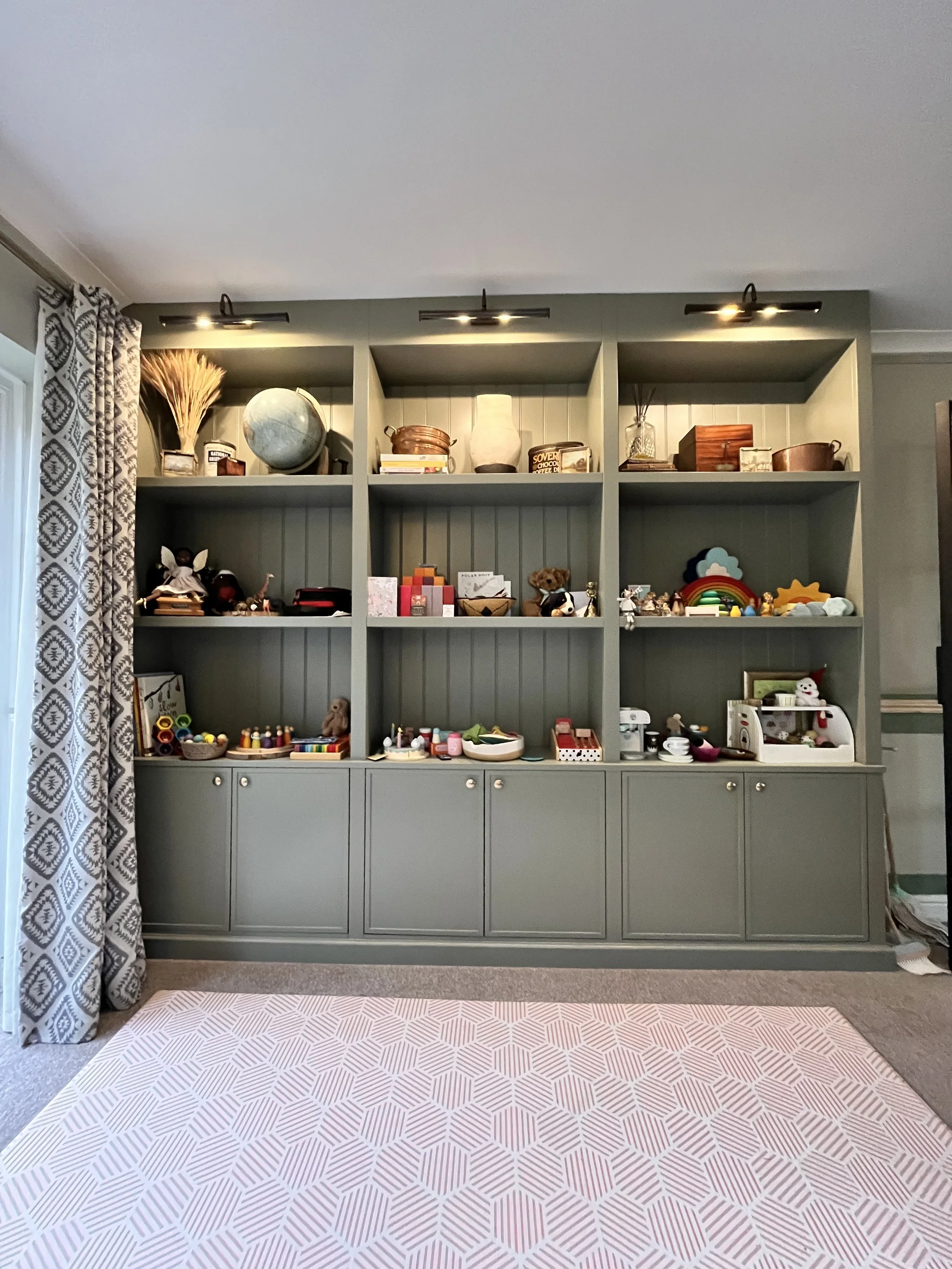 Gray built-in shelves filled with toys, books, and decorative items in a living room, with a pink patterned rug on the floor and patterned curtains on the left.