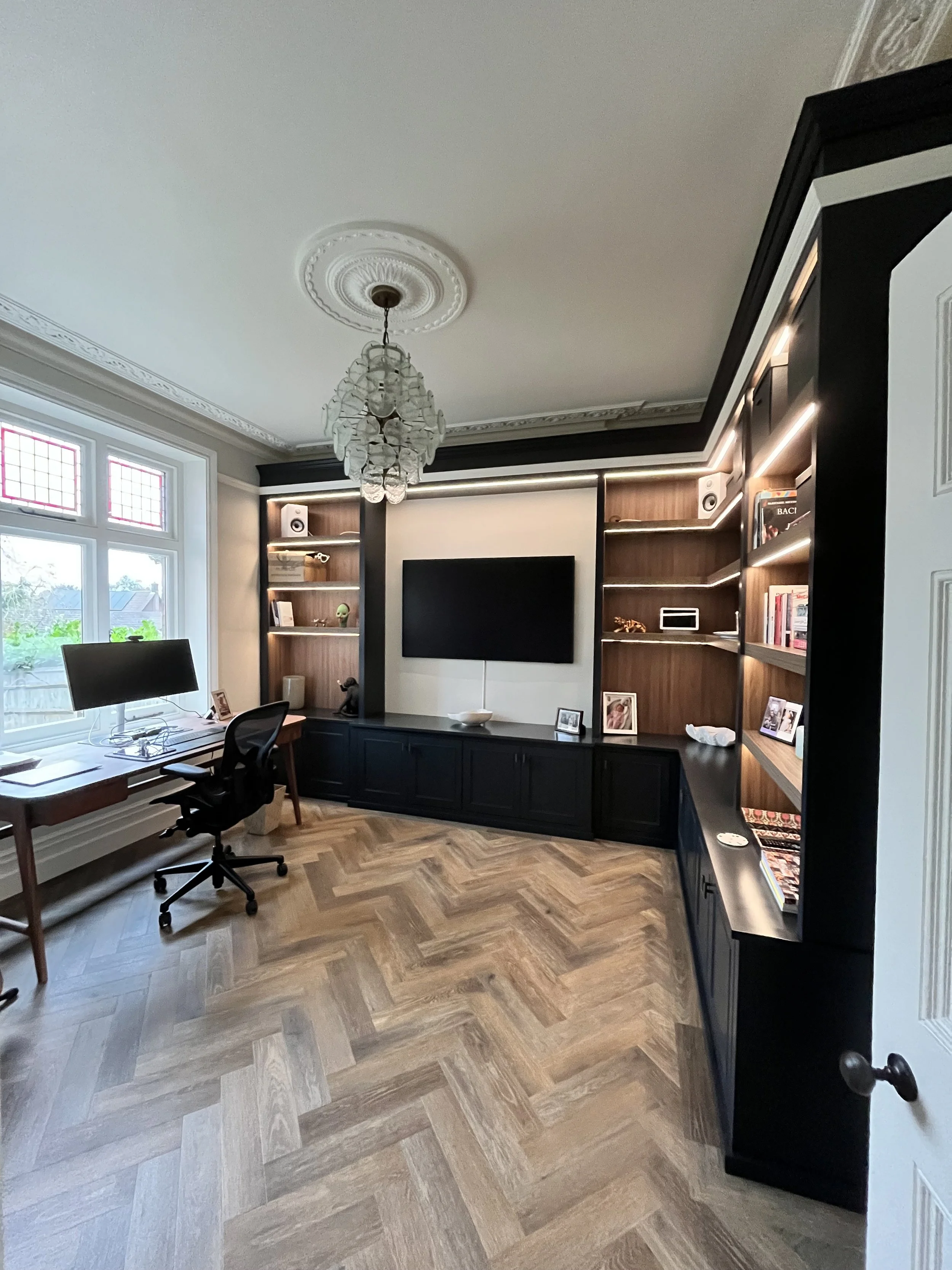 Home office with large window, black built-in shelves with books and decorations, wall-mounted TV, wooden floors, desk with computer, and chandelier lighting.