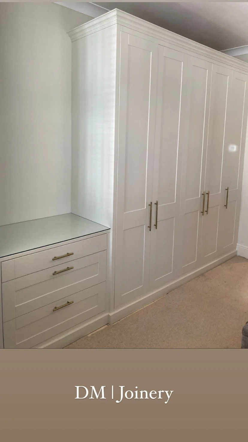 Built-in white wardrobe with gold handles in a room with beige carpet, and a small matching drawer unit with gold handles.