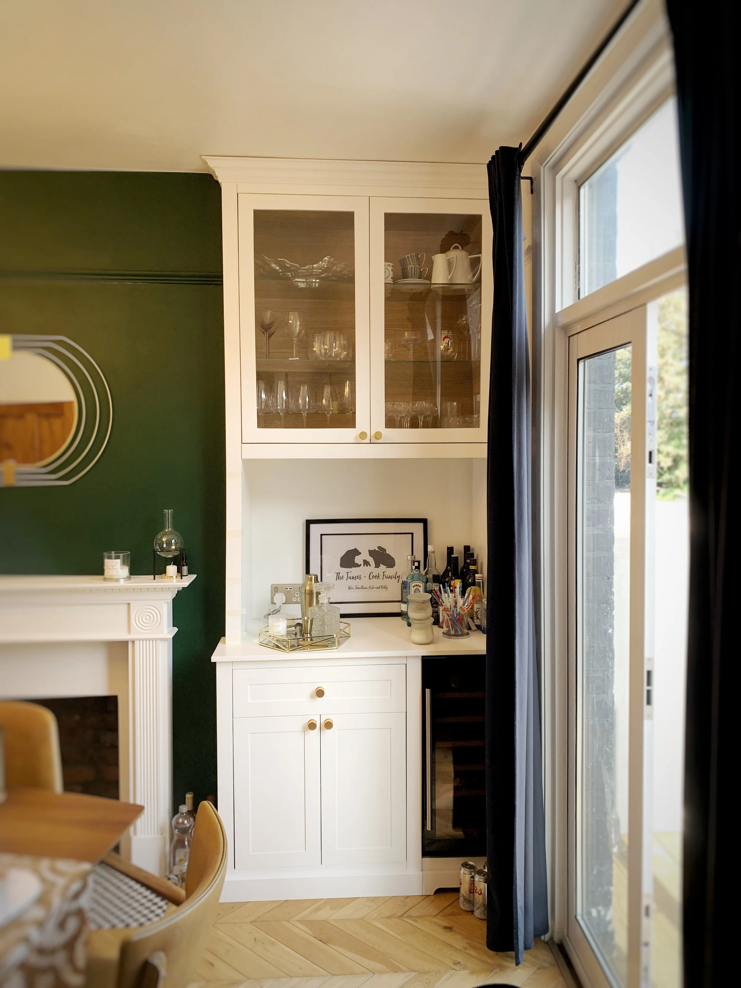 A white cabinet with glass doors displaying glassware, situated in a room with green and cream walls and a sliding glass door with dark curtains.