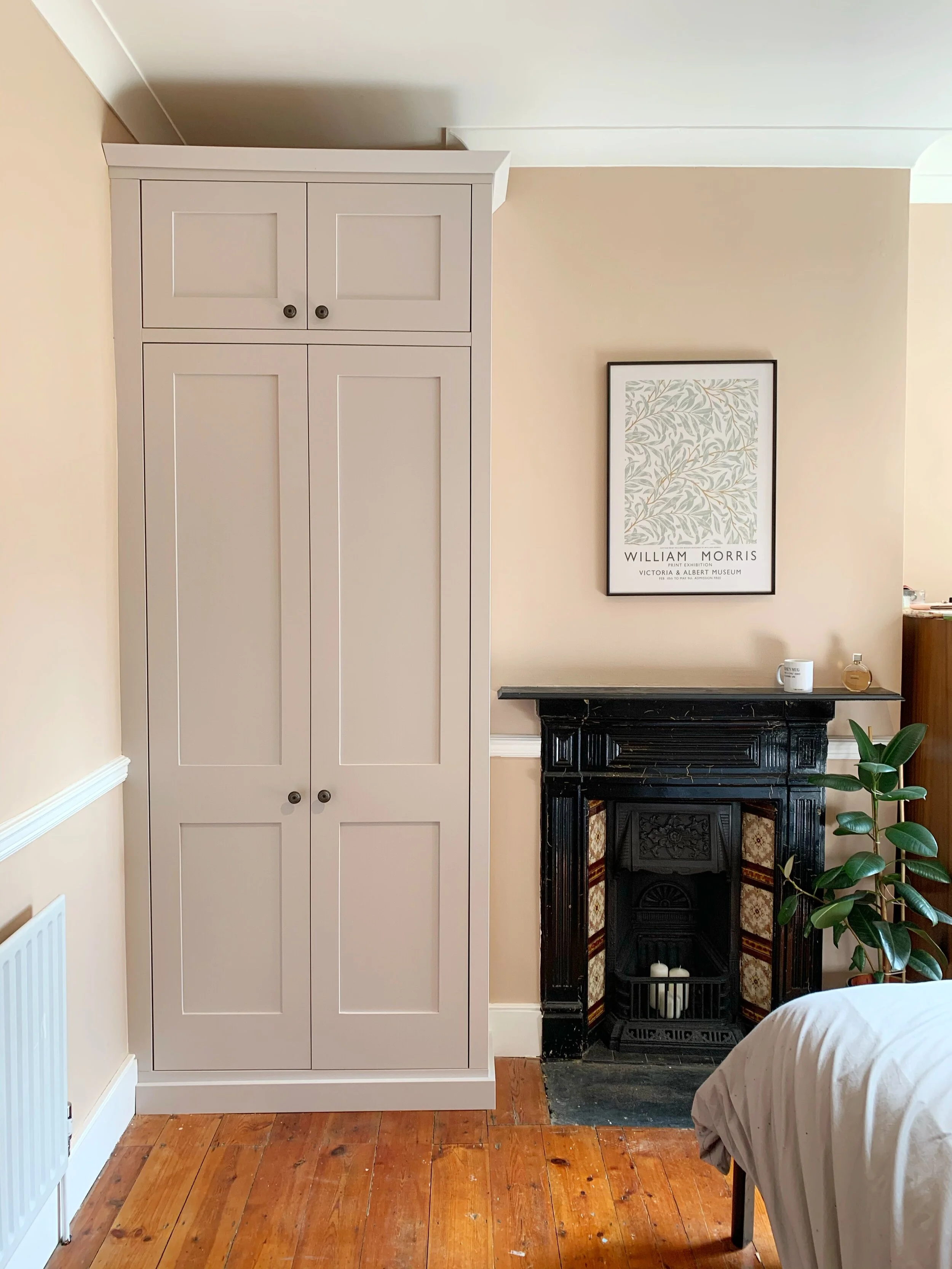 A bedroom corner with a beige wardrobe, a black vintage fireplace, a framed William Morris poster on the wall, a potted plant, part of a bed, and hardwood flooring.