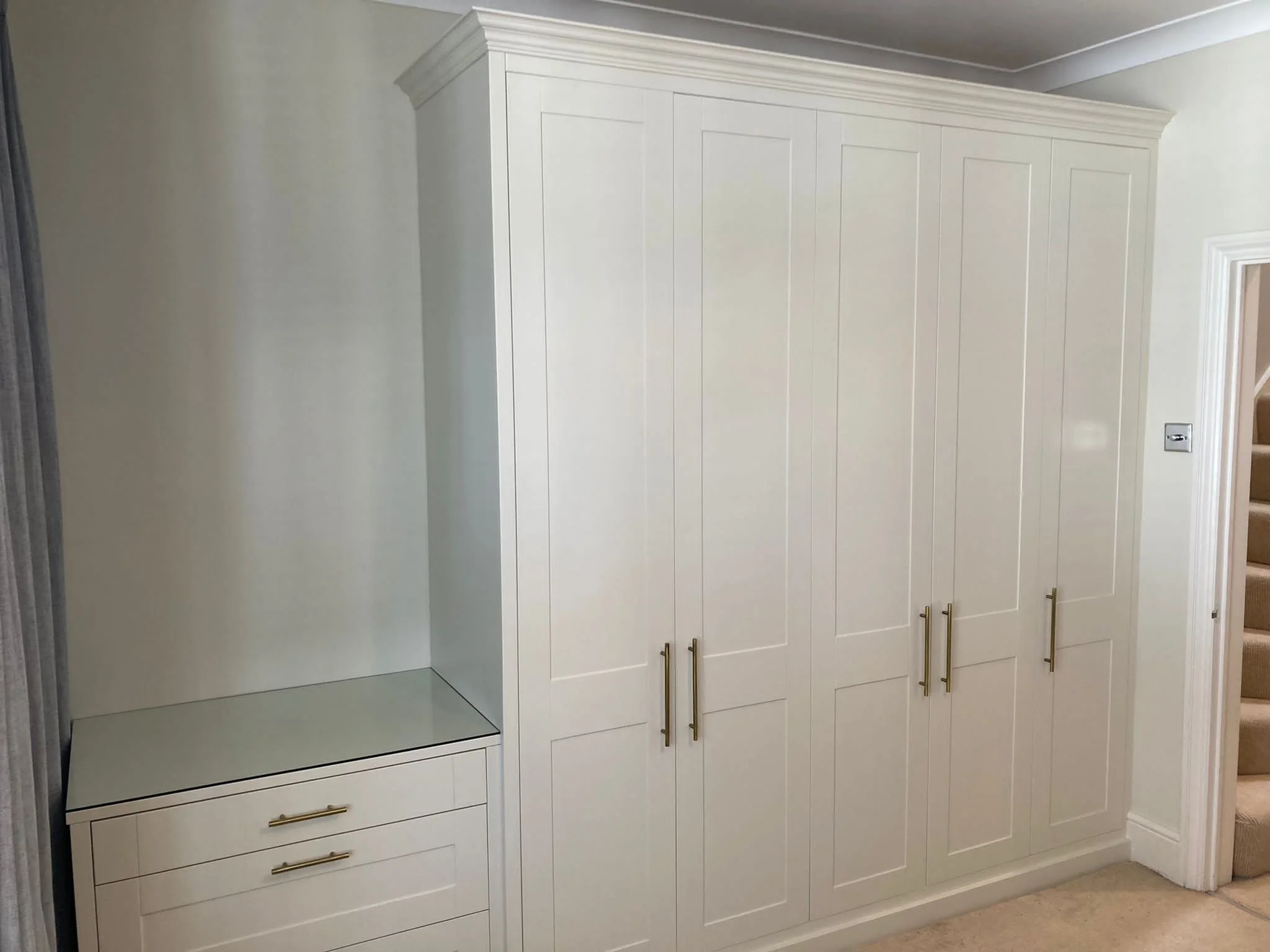 White built-in wardrobe with gold handles next to a small white dresser with gold handles, located in a room with beige carpet and stairs nearby.