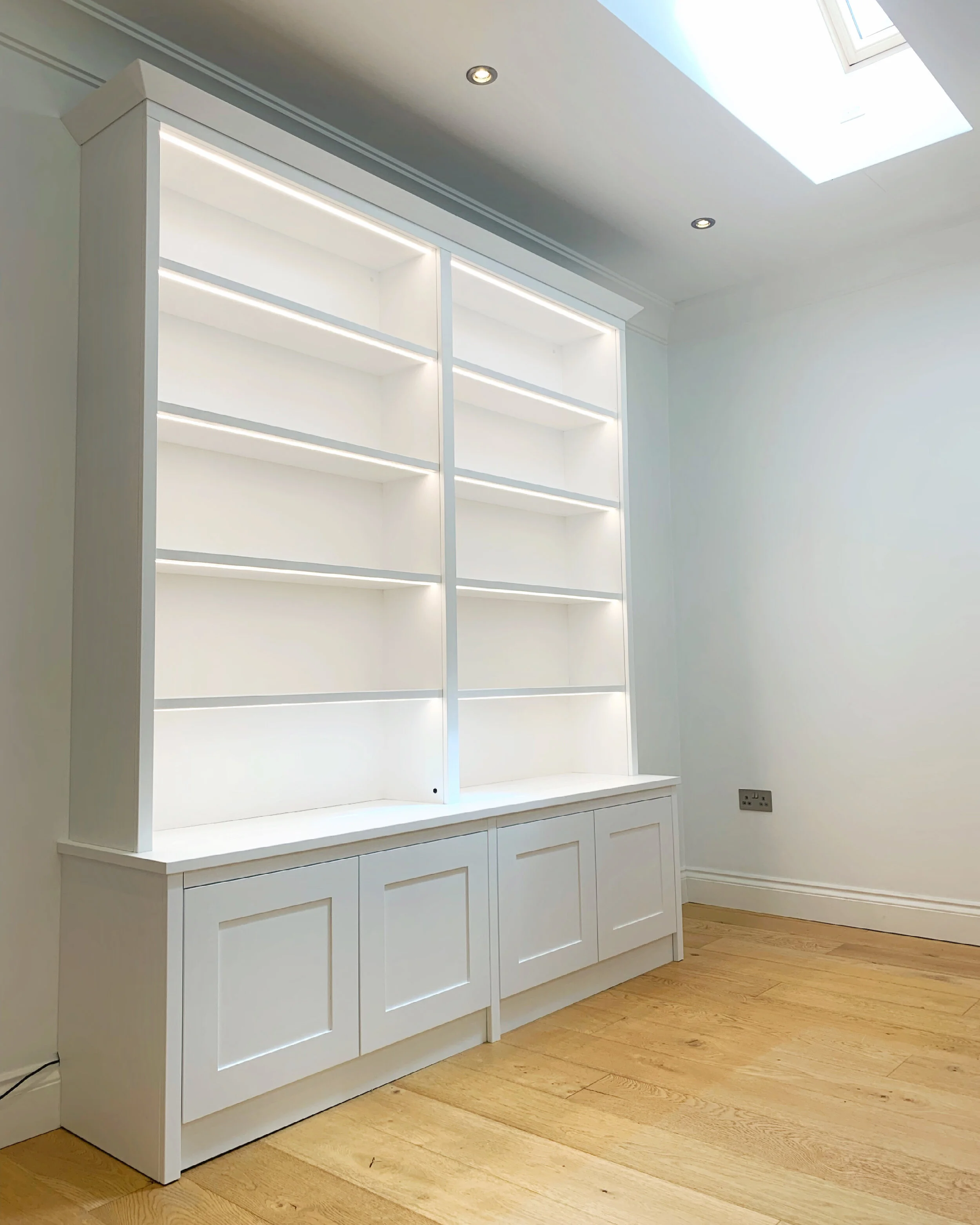White built-in bookshelf with open shelves and a closed cabinet base in a room with hardwood flooring and ceiling skylight.