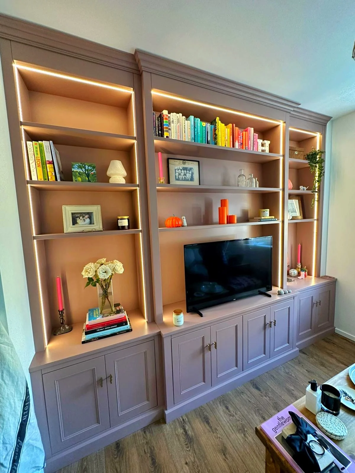 Pink built-in bookshelf with illuminated shelves, decorated with books, framed photos, candles, and decorative objects, and a TV in the center, in a living room.