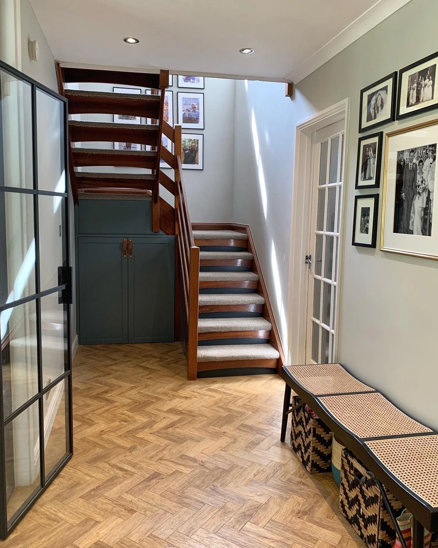 Entryway with wooden staircase, framed photographs on wall, glass door, and a small table with woven baskets underneath.
