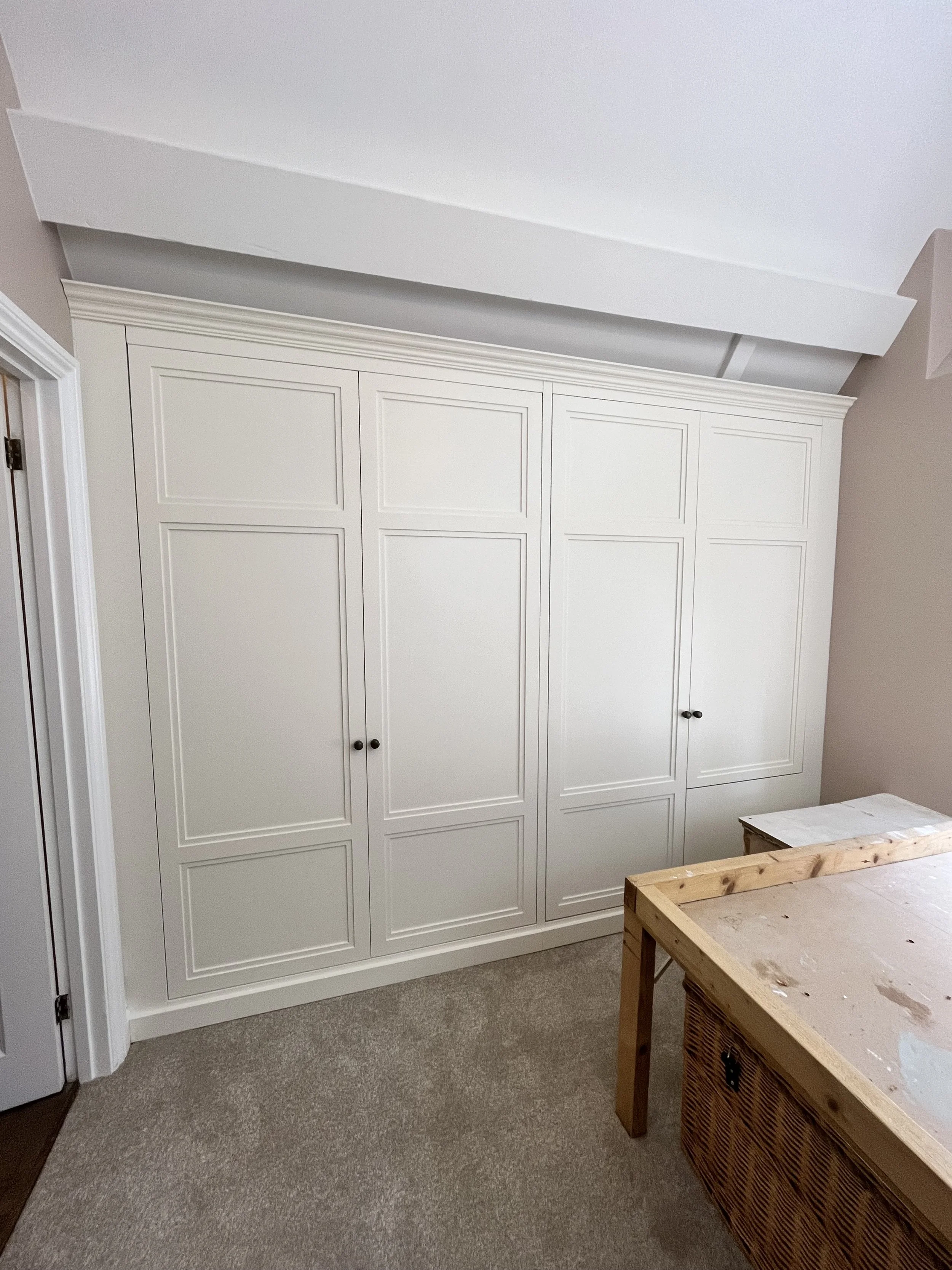 A large white built-in closet with paneled doors in a room under renovation. There is a wooden table or cabinet with a parchment top and a partially visible wicker basket below. The room has beige walls and a light brown carpet.