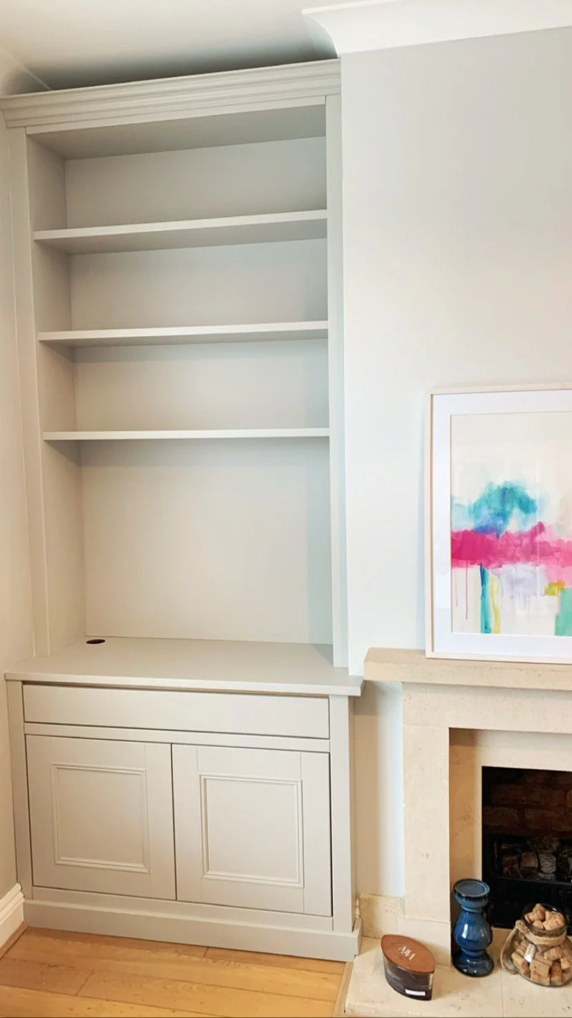 Empty built-in white bookshelf with two doors below, next to a fireplace with colorful abstract painting, candles, and decorative objects on the hearth.