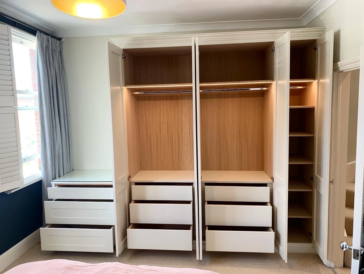 Empty white wardrobe with compartments, drawers, and shelves in a bedroom with a window and curtains.
