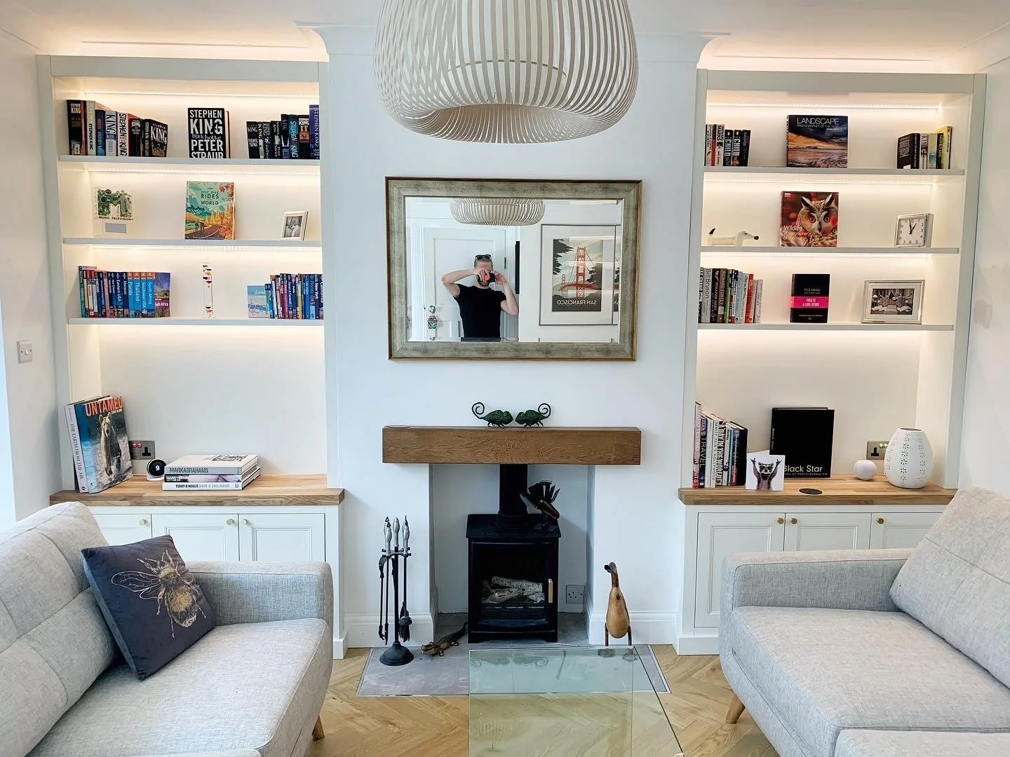 Living room with built-in bookshelves, a mirror above a small fireplace, and two beige sofas facing each other. The bookshelves are lit and contain books and decorative items. There is a ceiling light fixture and a person taking a photo in the mirror