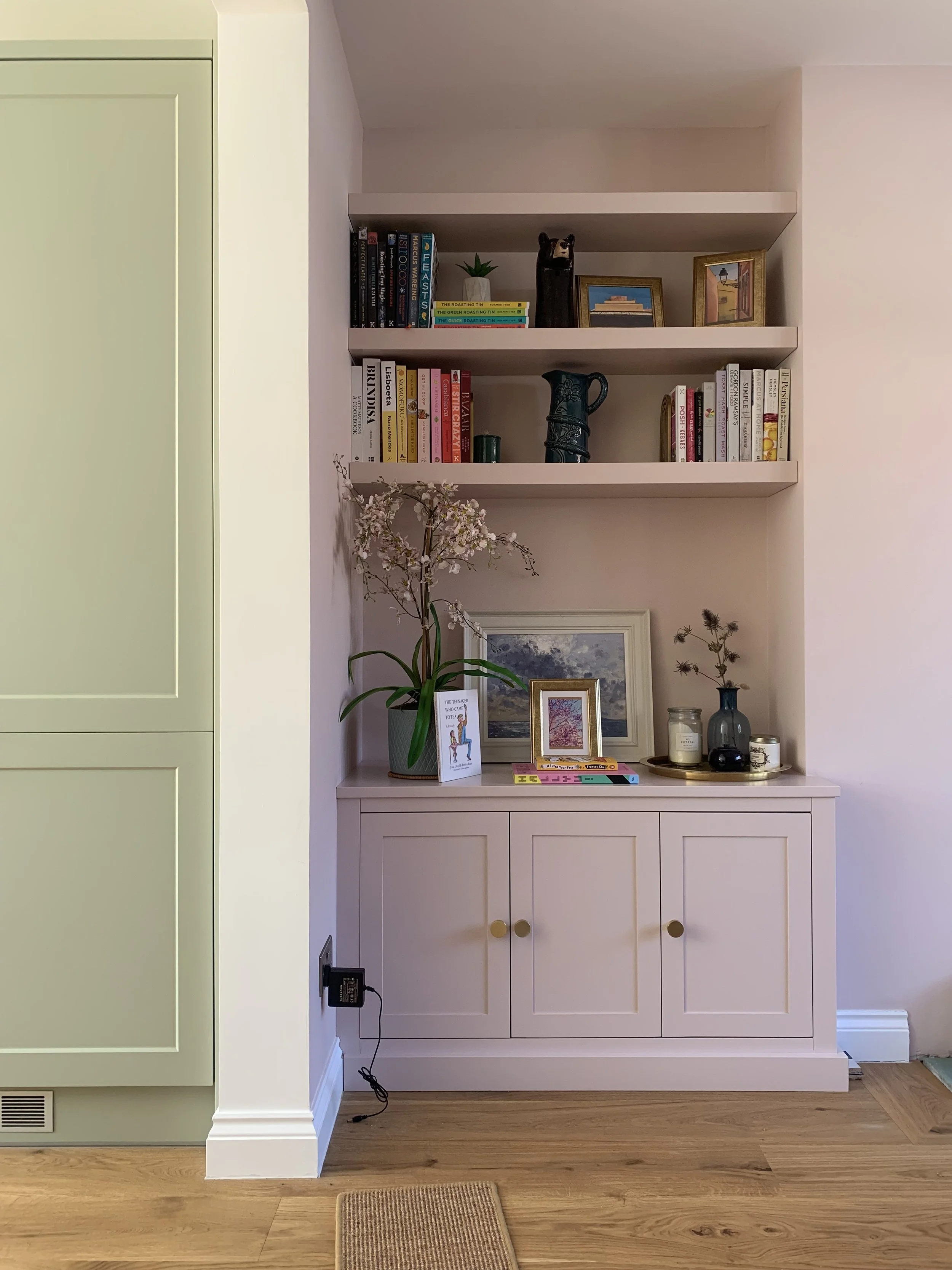 A white built-in bookshelf with three shelves filled with books, decorative items, and framed pictures. A potted orchid with pink blossoms is on top of a white cabinet below the shelves, which has three doors with round gold knobs. There are various 