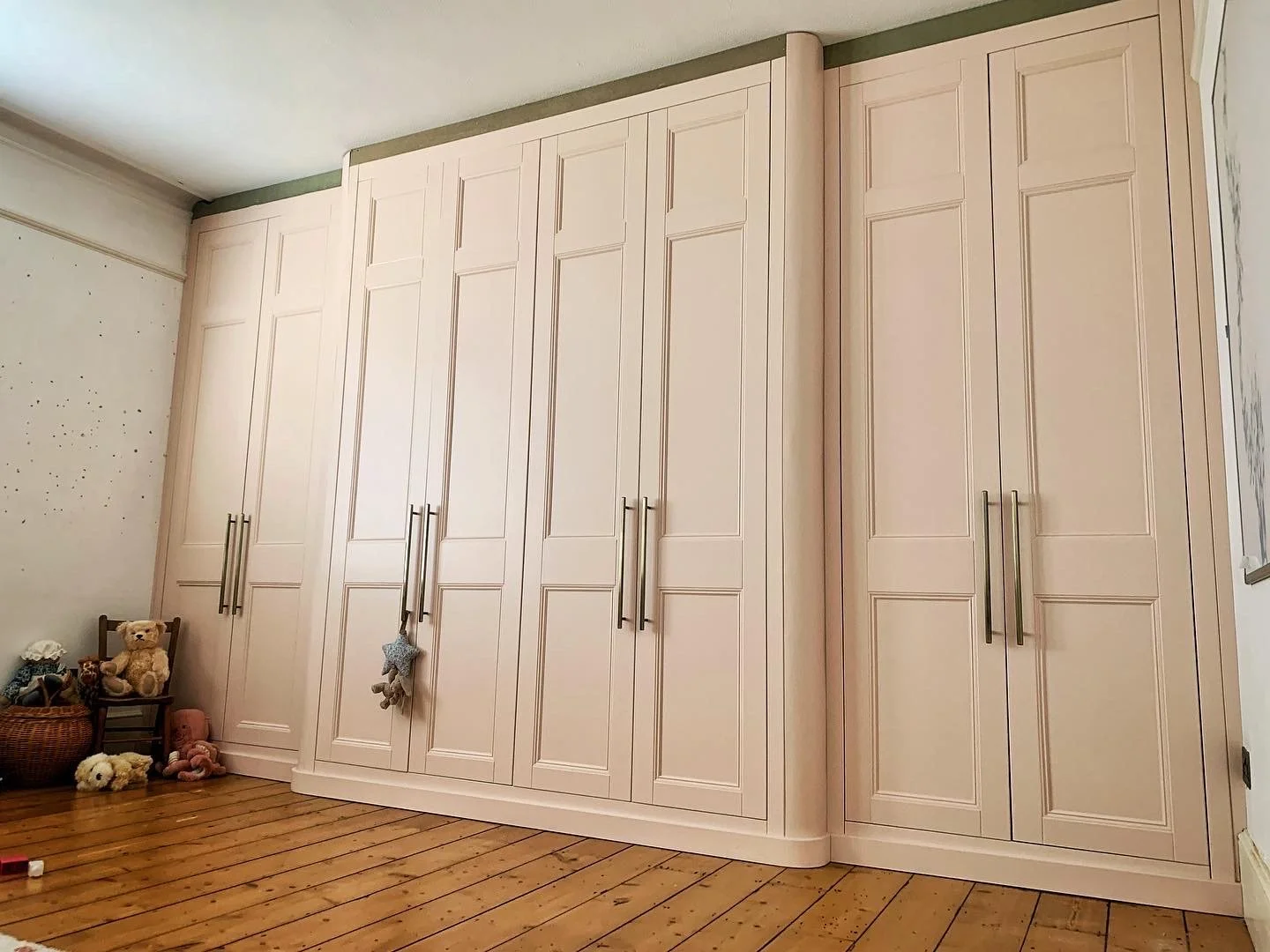 Large white built-in wardrobe with metal handles in a room with wooden floor and toys, including teddy bears, on the floor.