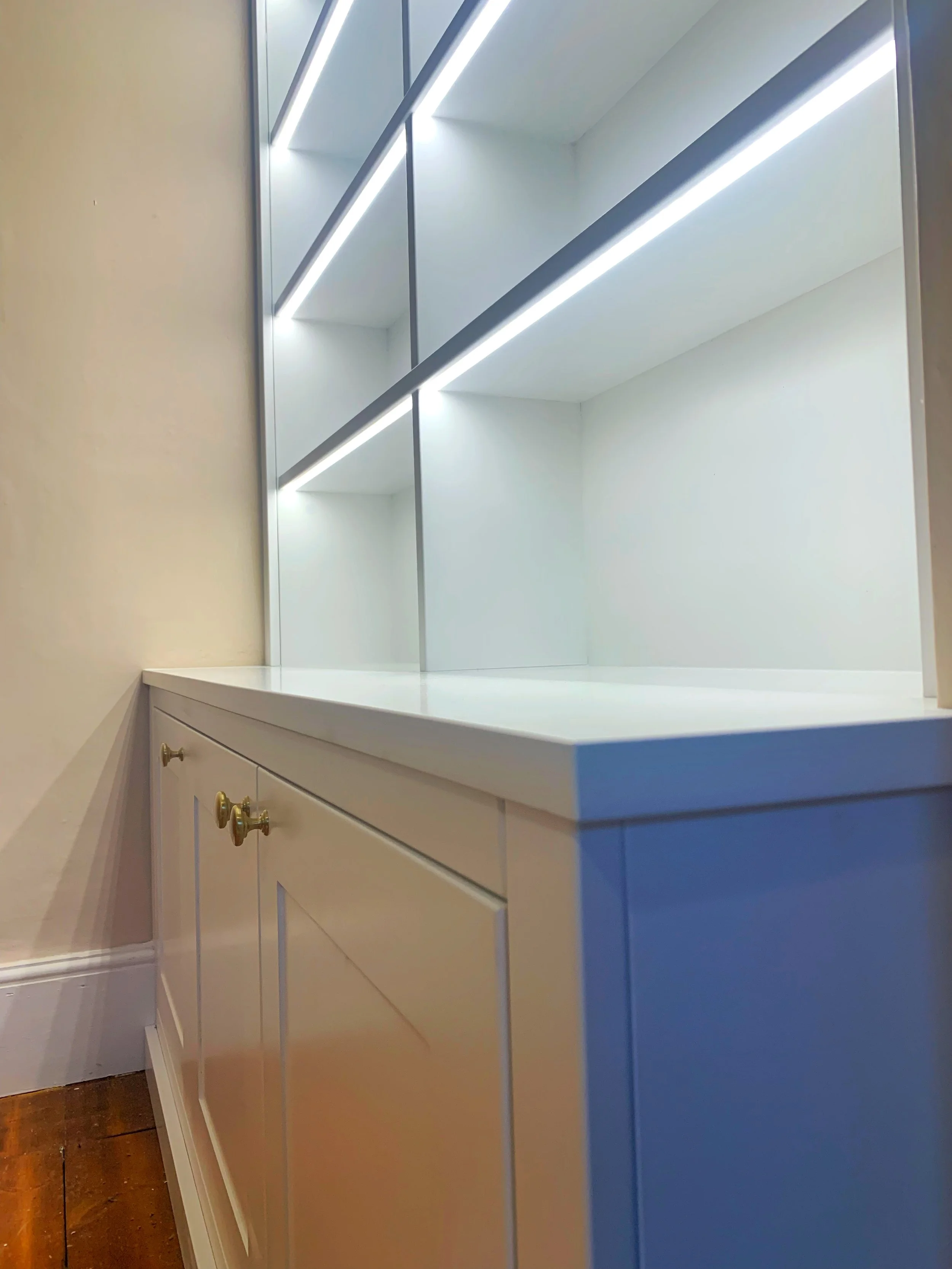 A white modern cabinet with gold knobs and open shelving illuminated with built-in LED lighting.