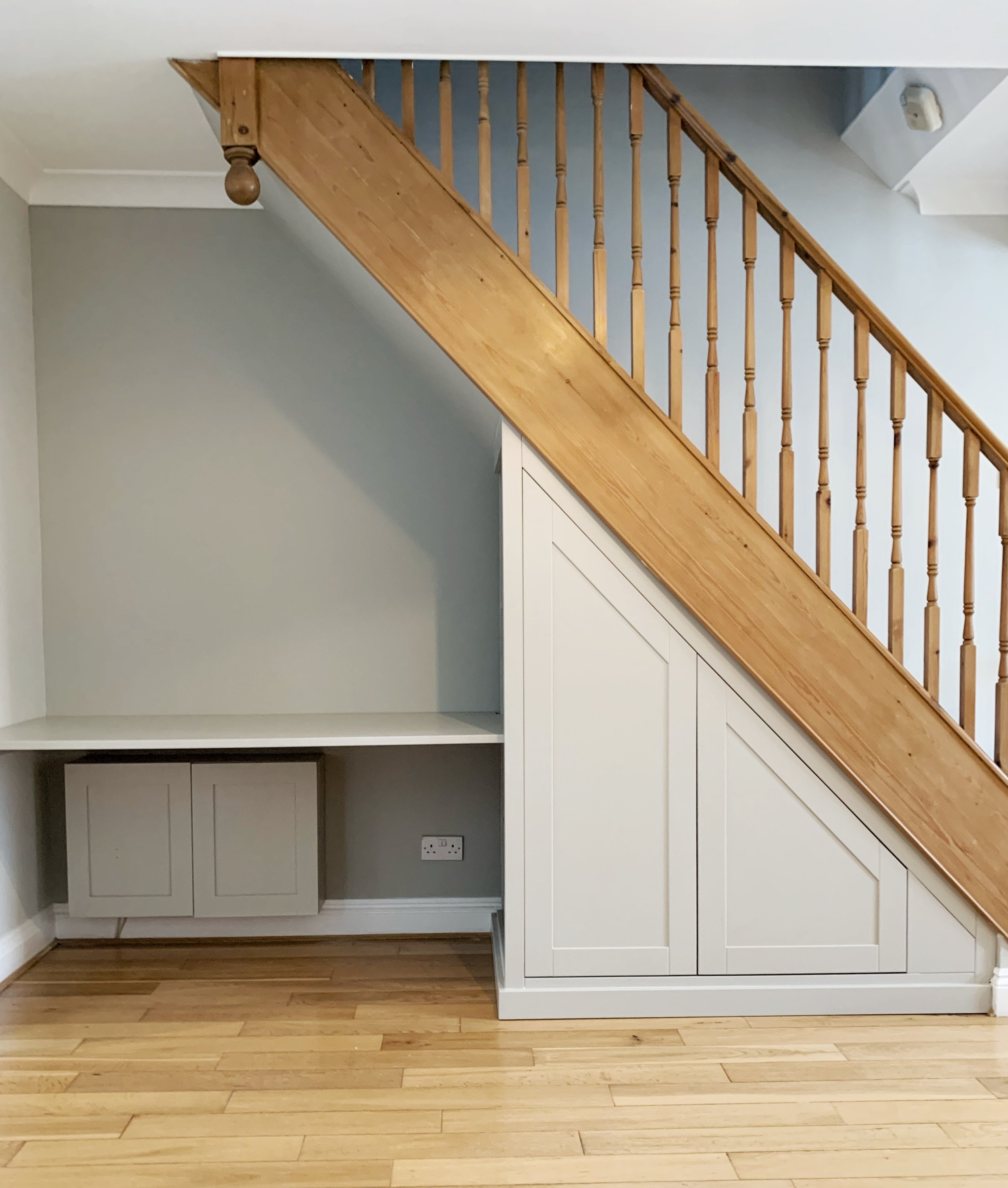 Interior of a room with a staircase overhead and built-in storage cabinets underneath, featuring wood flooring and a gray wall.