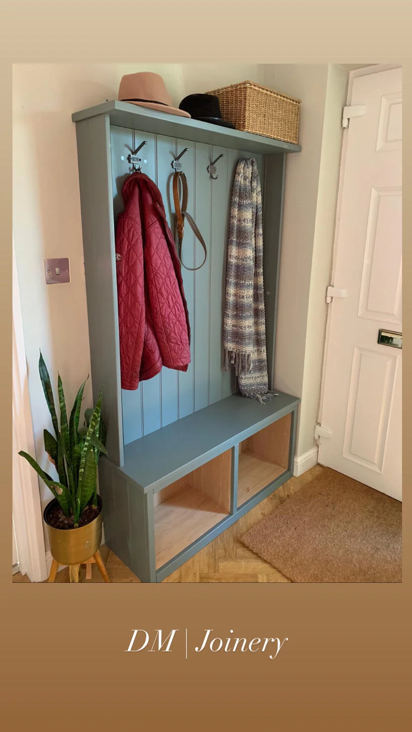 A blue wooden coat rack with hooks holding a red quilted jacket, a brown leather strap, and a multicolored plaid scarf. The top shelf holds hats and a wicker basket, and the bottom has open cubbies. Next to the coat rack is a potted snake plant on a 