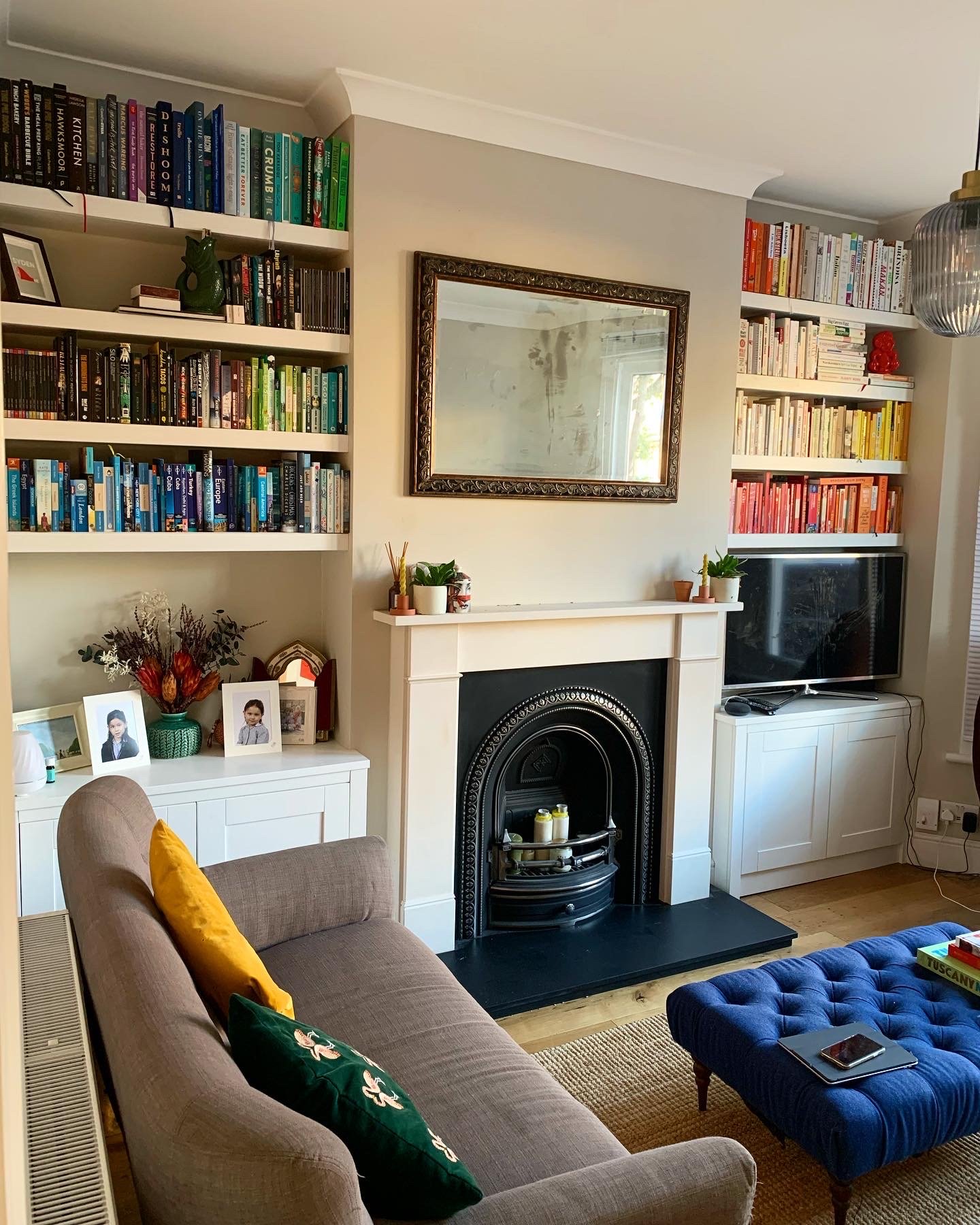 Living room with built-in bookshelves, a mirror above a fireplace, a beige sofa with yellow and green cushions, a blue tufted ottoman with a book and phone on top, and a flat-screen TV next to the fireplace.