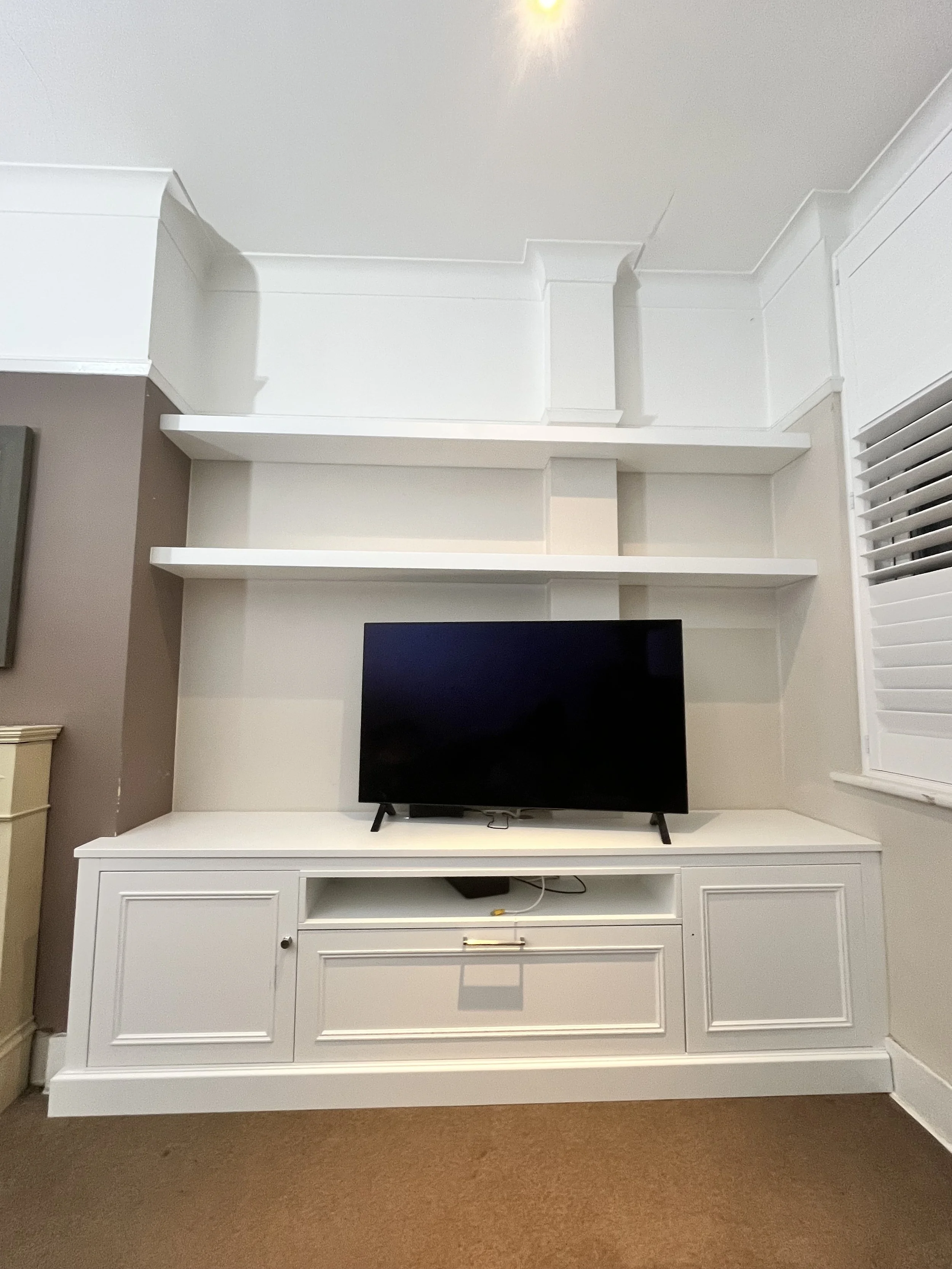 White entertainment center with a flat-screen TV, open shelves, and a window with blinds on the right.