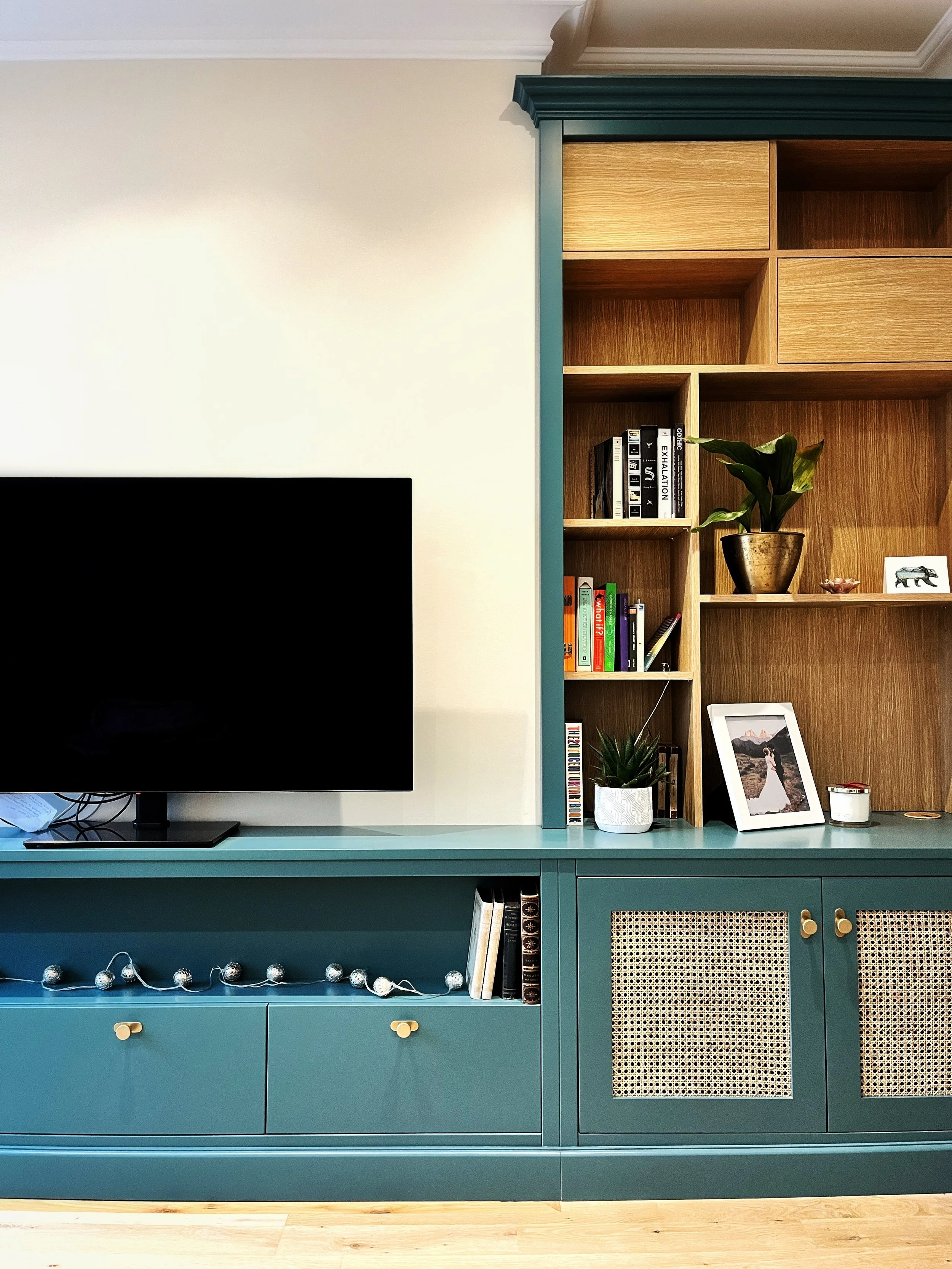 A teal TV stand with books and decorative items, a flat-screen TV, and a wooden bookshelf with potted plants and framed photos.