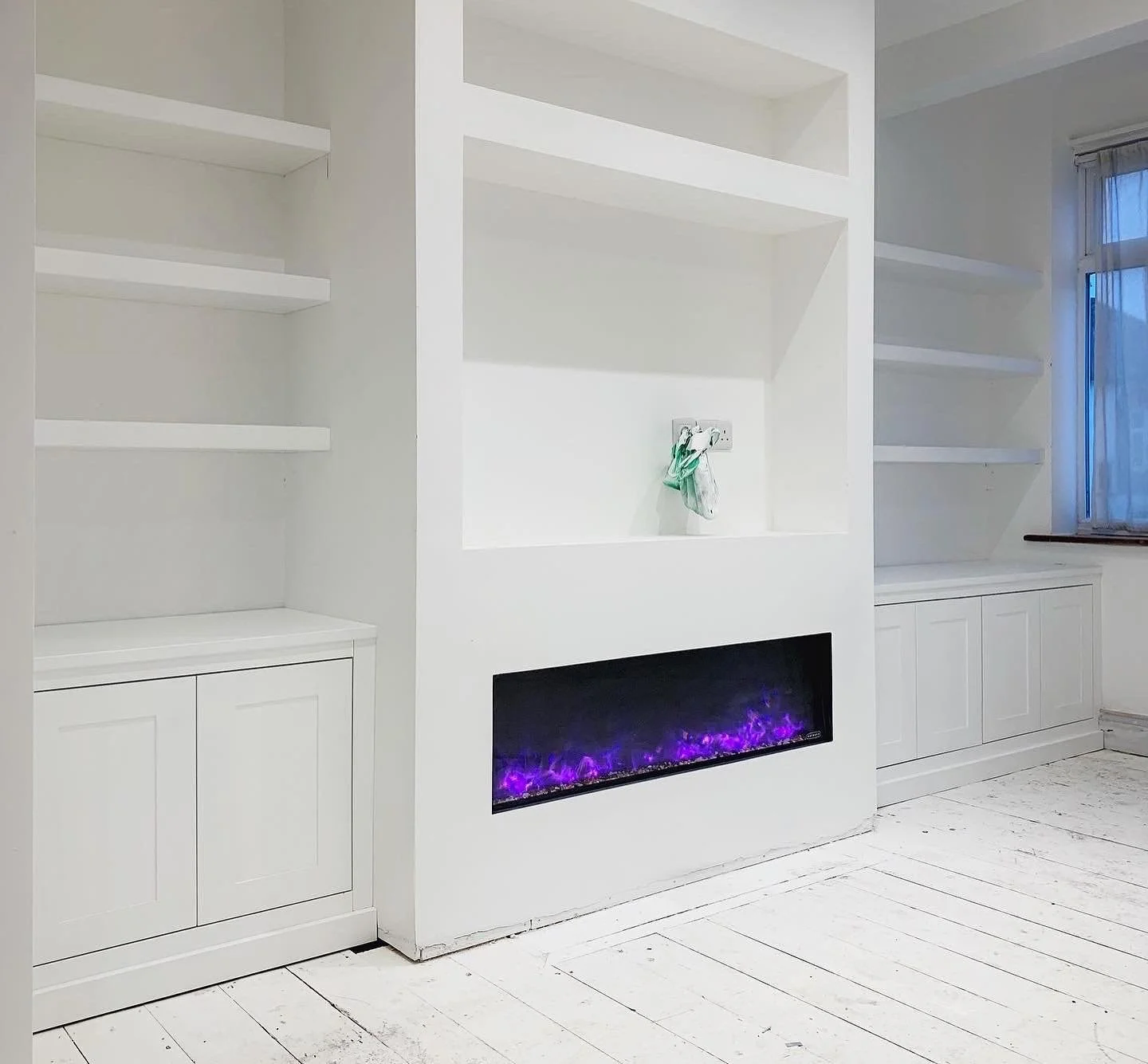 Empty white built-in cabinets and shelves in a room with a modern electric fireplace with purple flames, and a window with sheer curtains.