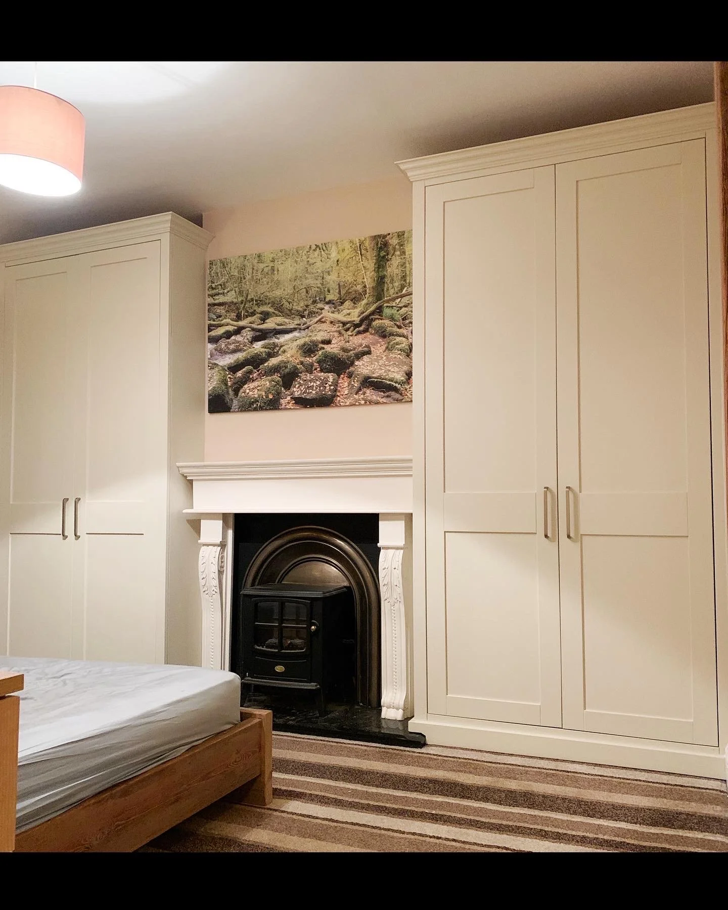Bedroom with white wardrobe, a fireplace with decorative white columns, a nature-themed painting above the fireplace, a bed with a mattress, and striped carpet.