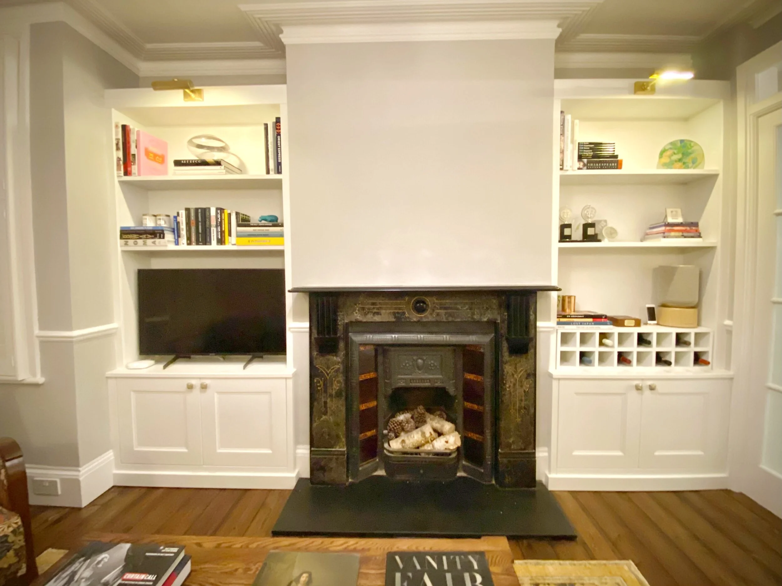 A living room with a fireplace flanked by built-in white bookshelves, a flat-screen TV, and various books and decorative items. The fireplace has a black marble mantel and logs inside, and there are hardwood floors.