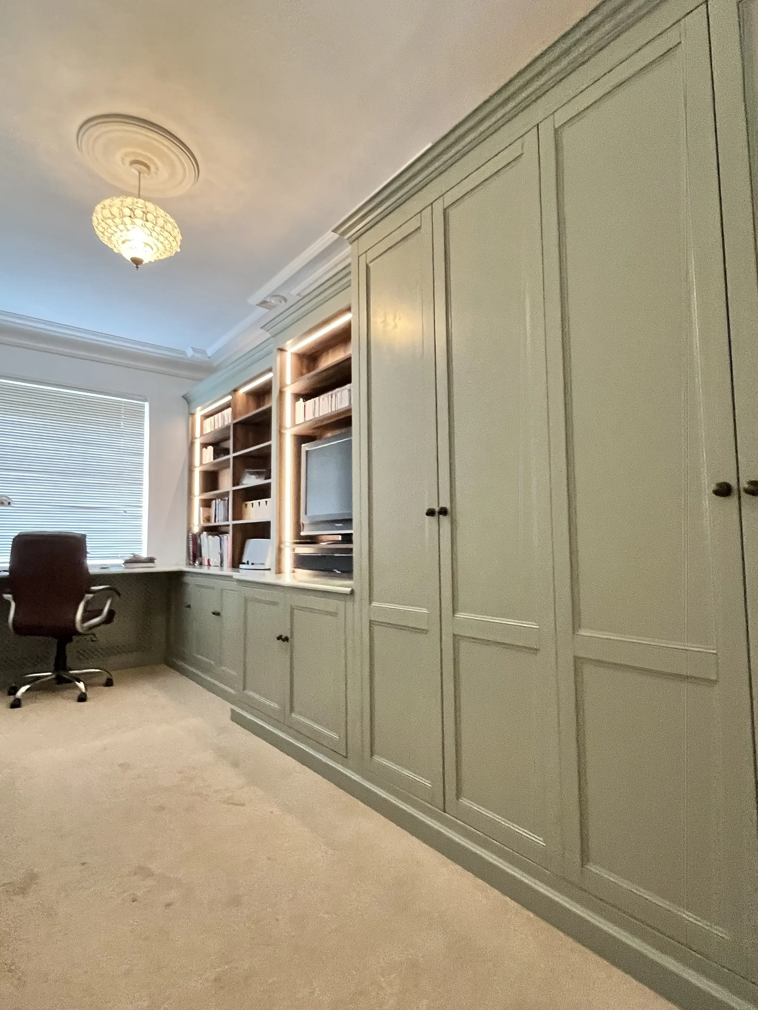 Home office with built-in grey cabinets, a wall-mounted TV, bookshelves with books, a desk with a black office chair, a window with blinds, and a ceiling light fixture.