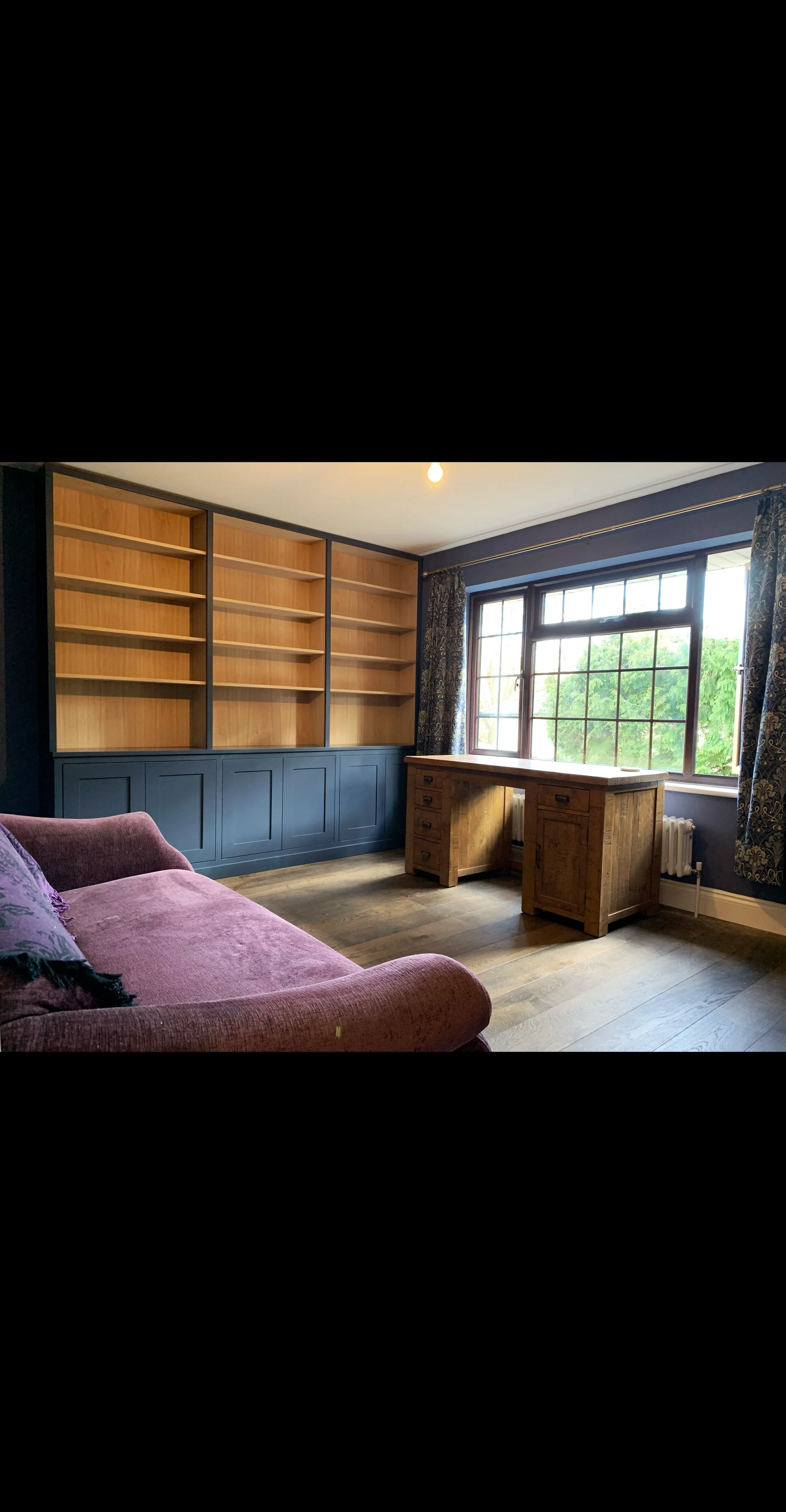 Room with built-in wooden bookshelves, a wooden desk, a purple sofa, and large window with curtains showing green trees outside.