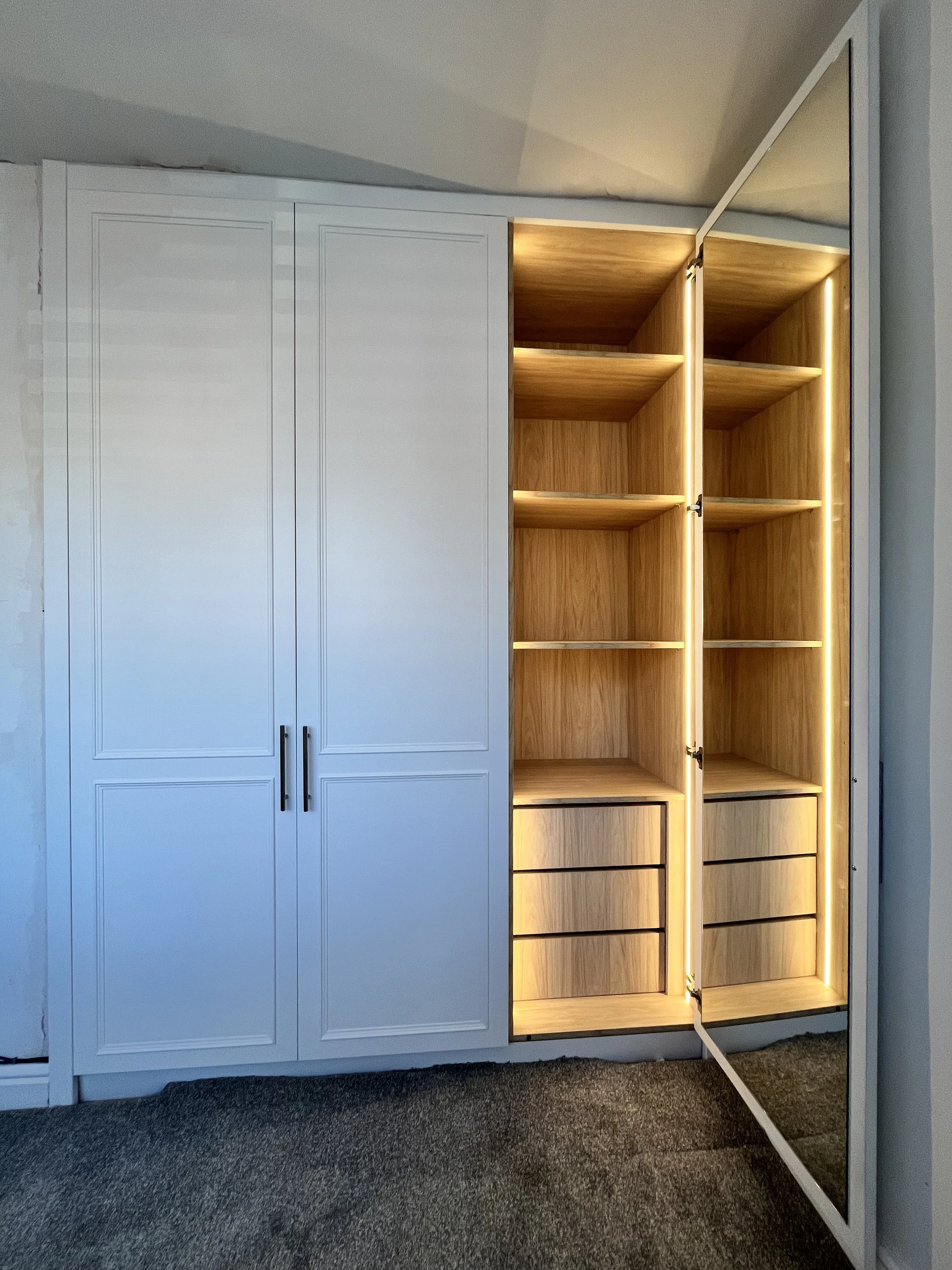 A white wardrobe with a partially open mirror door revealing wooden shelves and drawers inside.