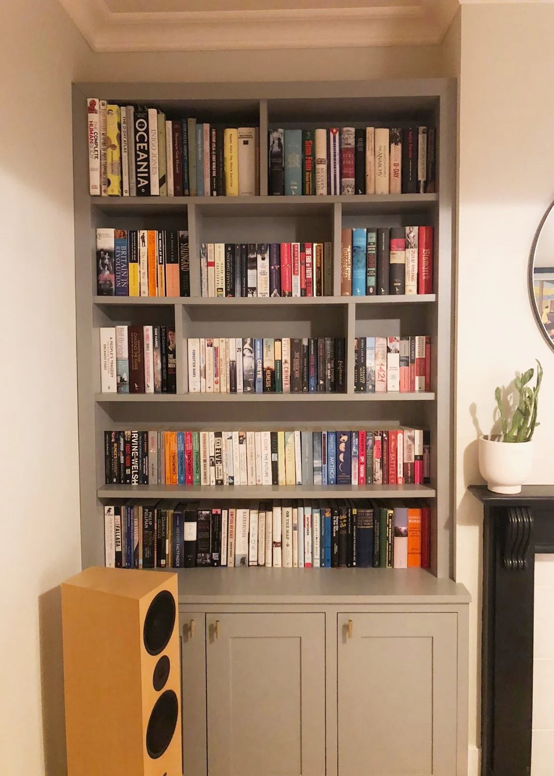 A tall grey bookshelf filled with a variety of books, positioned against a beige wall. To the right of the bookshelf is a mirror and a potted cactus on a black fireplace mantel. To the left, there is a yellow speaker with black speaker openings.