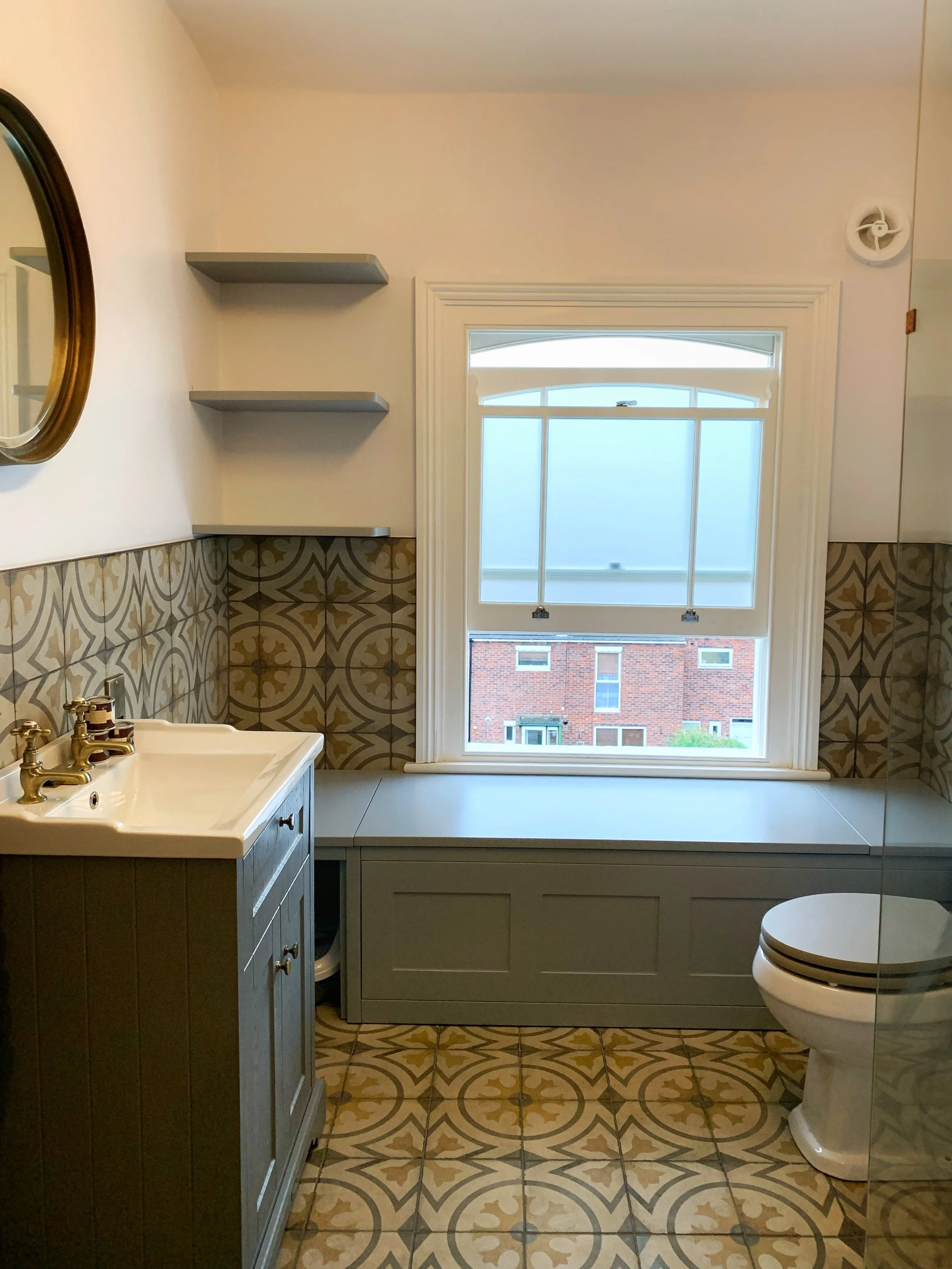 Bathroom with patterned floor tiles, a vanity with a sink on the left, a window with frosted glass in the center, and a toilet on the right. Two gray shelves are mounted on the wall above the vanity.