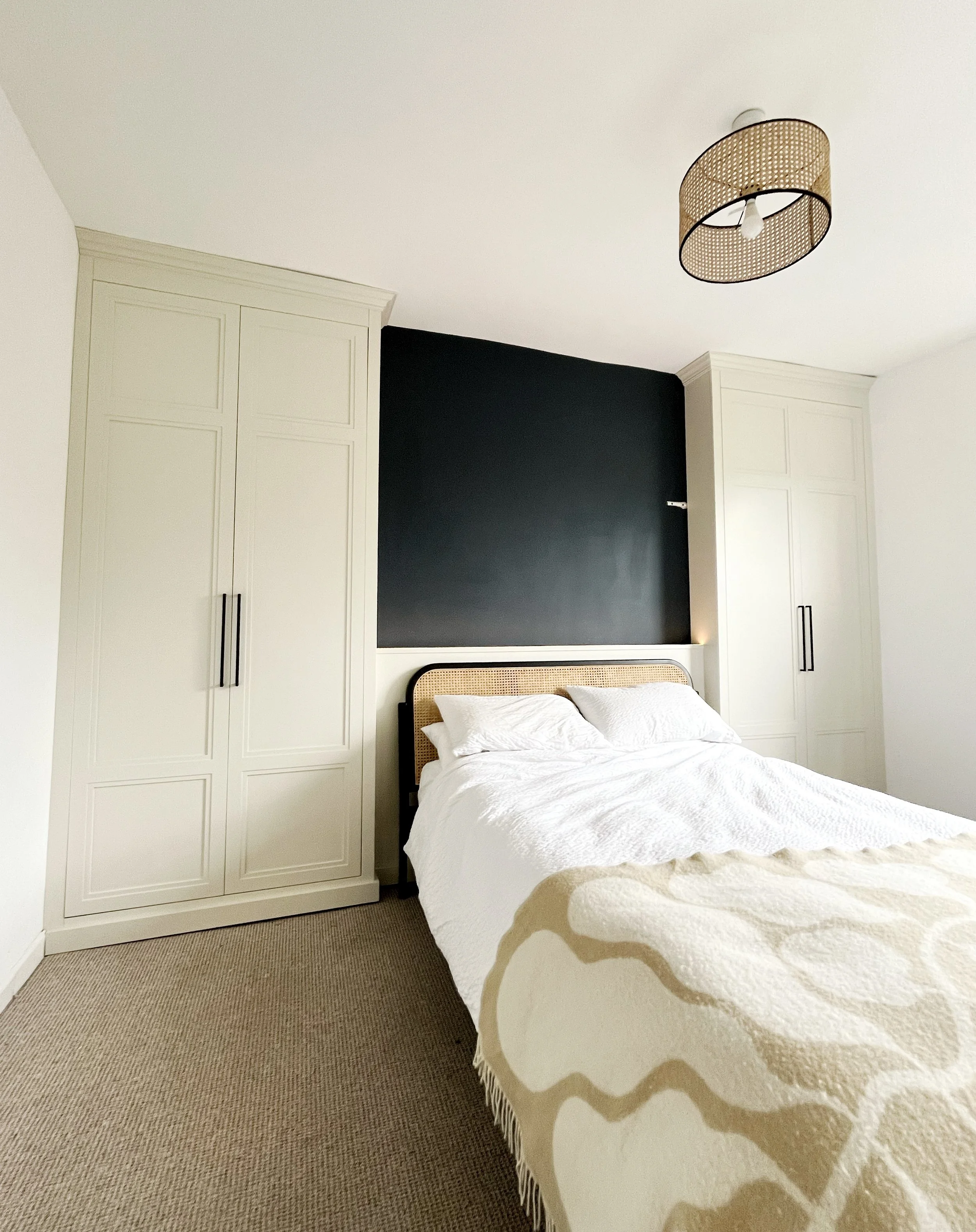 Modern bedroom with a bed that has a rattan headboard, white bedding, and black accents. Flanking the bed are built-in white wardrobes and a black chalkboard wall behind the bed. Ceiling light fixture with a woven rattan shade.