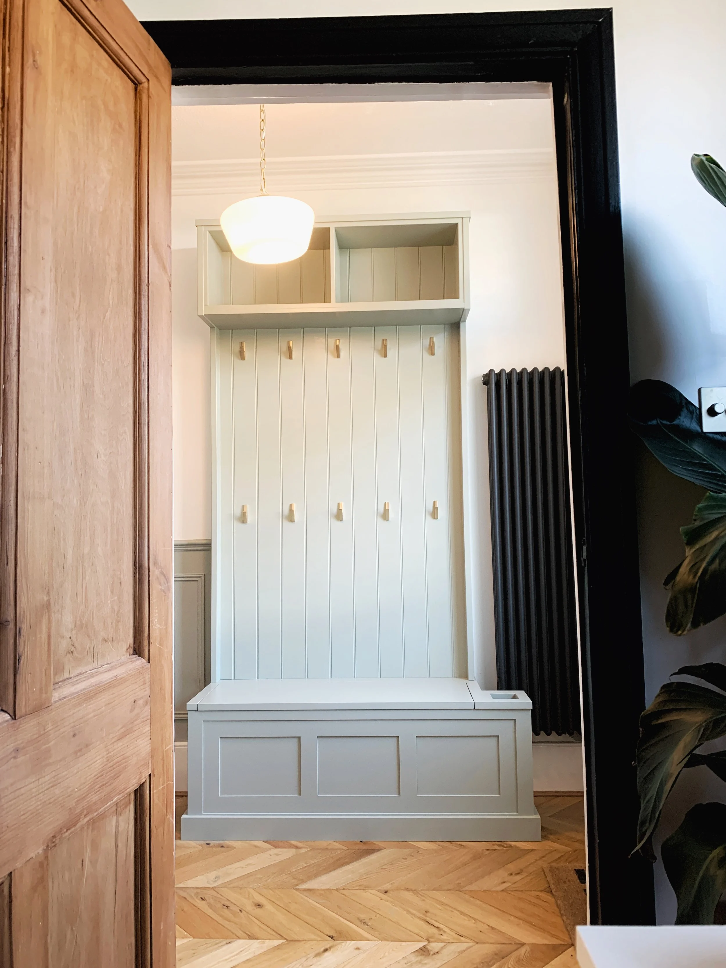 A white hall tree with hooks and storage bench, located in an entryway with hardwood flooring and a white wall. A black radiator and a plant are also visible.