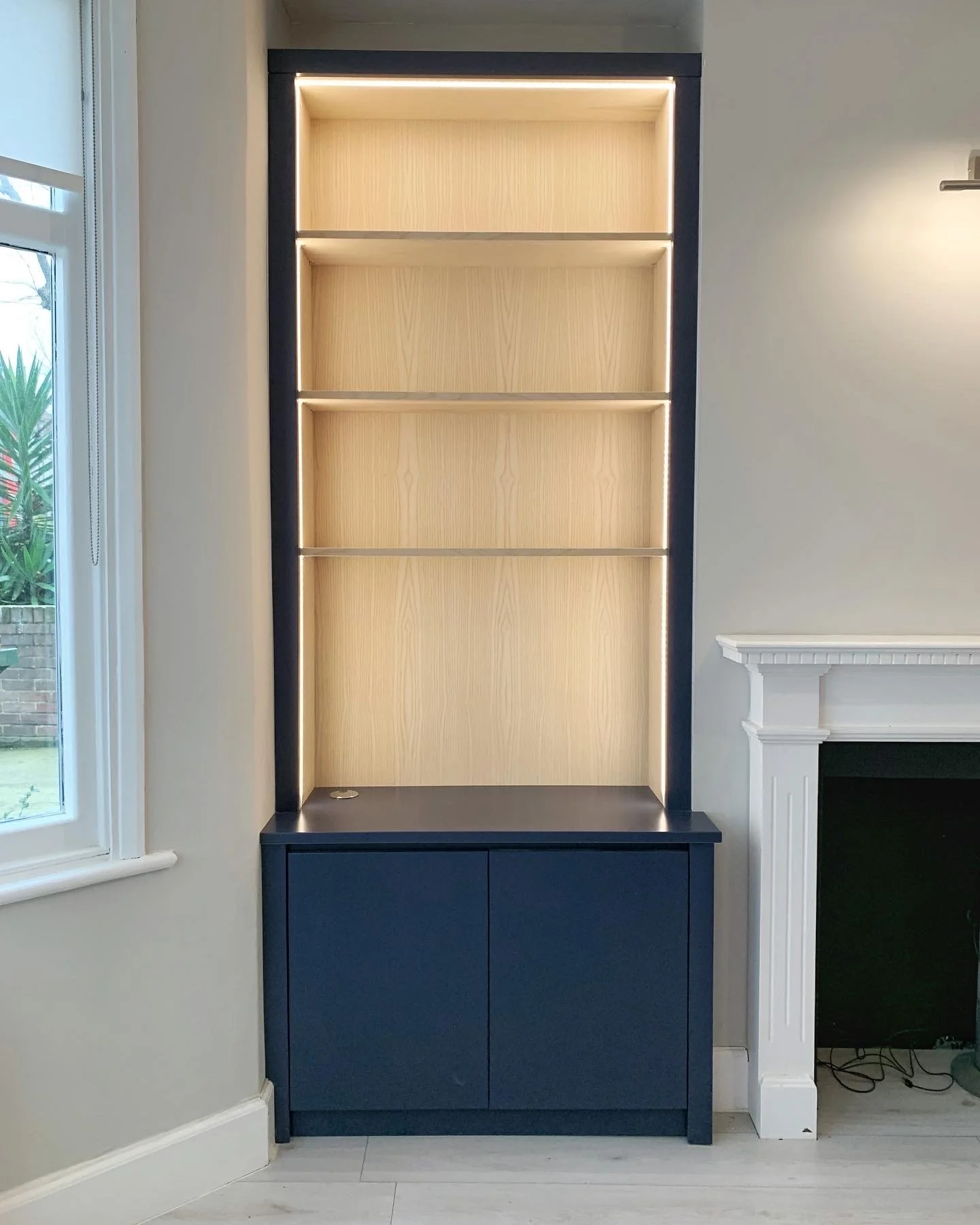 Empty built-in bookshelf with light wood shelves and a dark frame, positioned between a window and a fireplace in a living room.