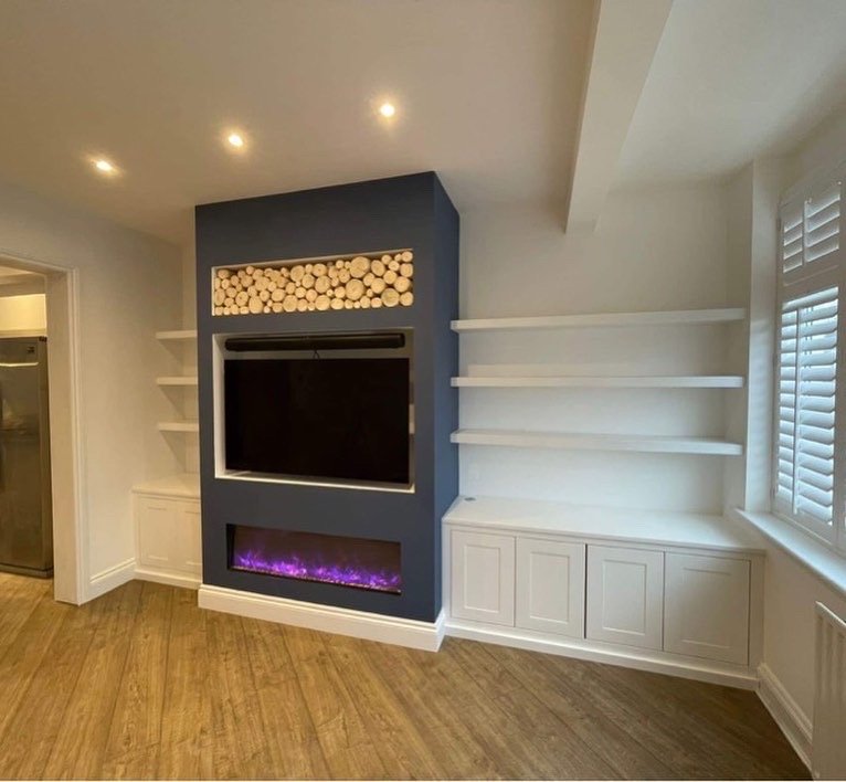 Living room with white shelves, built-in electric fireplace, and a dark blue accent wall with wood logs stored above the fireplace. Hardwood flooring and a window with white shutters are also visible.