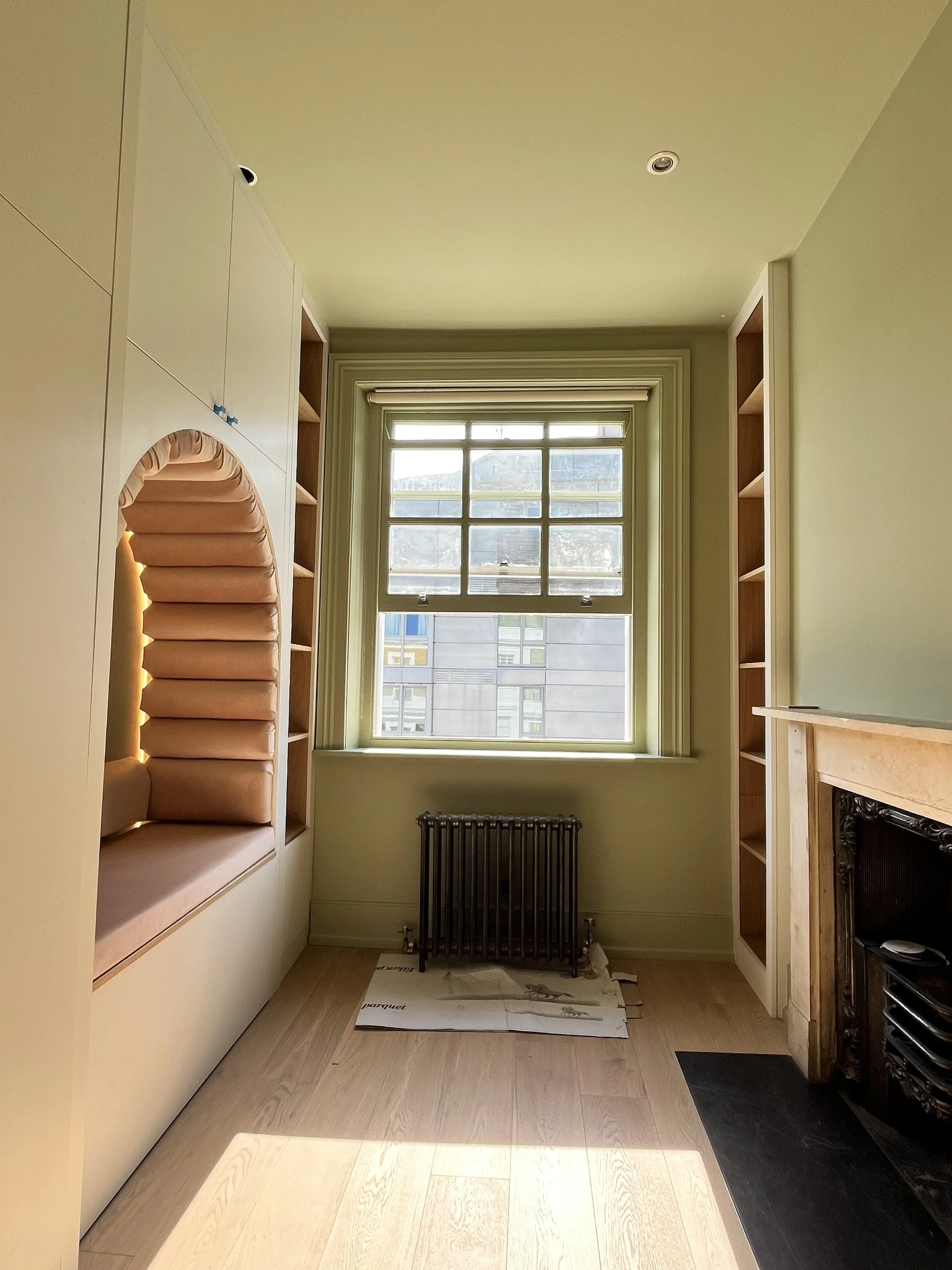 Empty room with built-in shelves on both sides, a window with multiple panes, a radiator below the window, and a fireplace with a decorative mantle on the right.