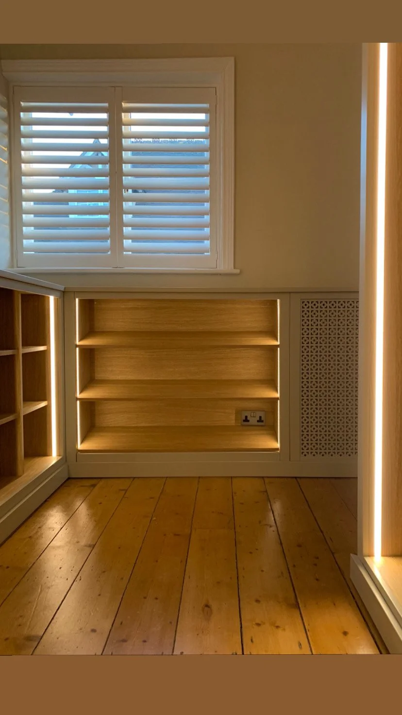 Empty built-in wooden shelves in a room with a window with white shutters, yellow walls, and wooden flooring.