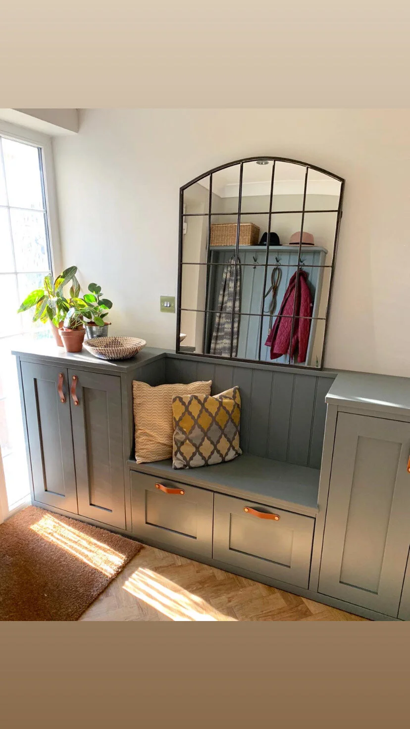 A gray built-in bench with cushions, positioned below a large decorative mirror with a grid pattern. To the left, a windowsill holds potted plants and a woven bowl. The area is illuminated by natural light.