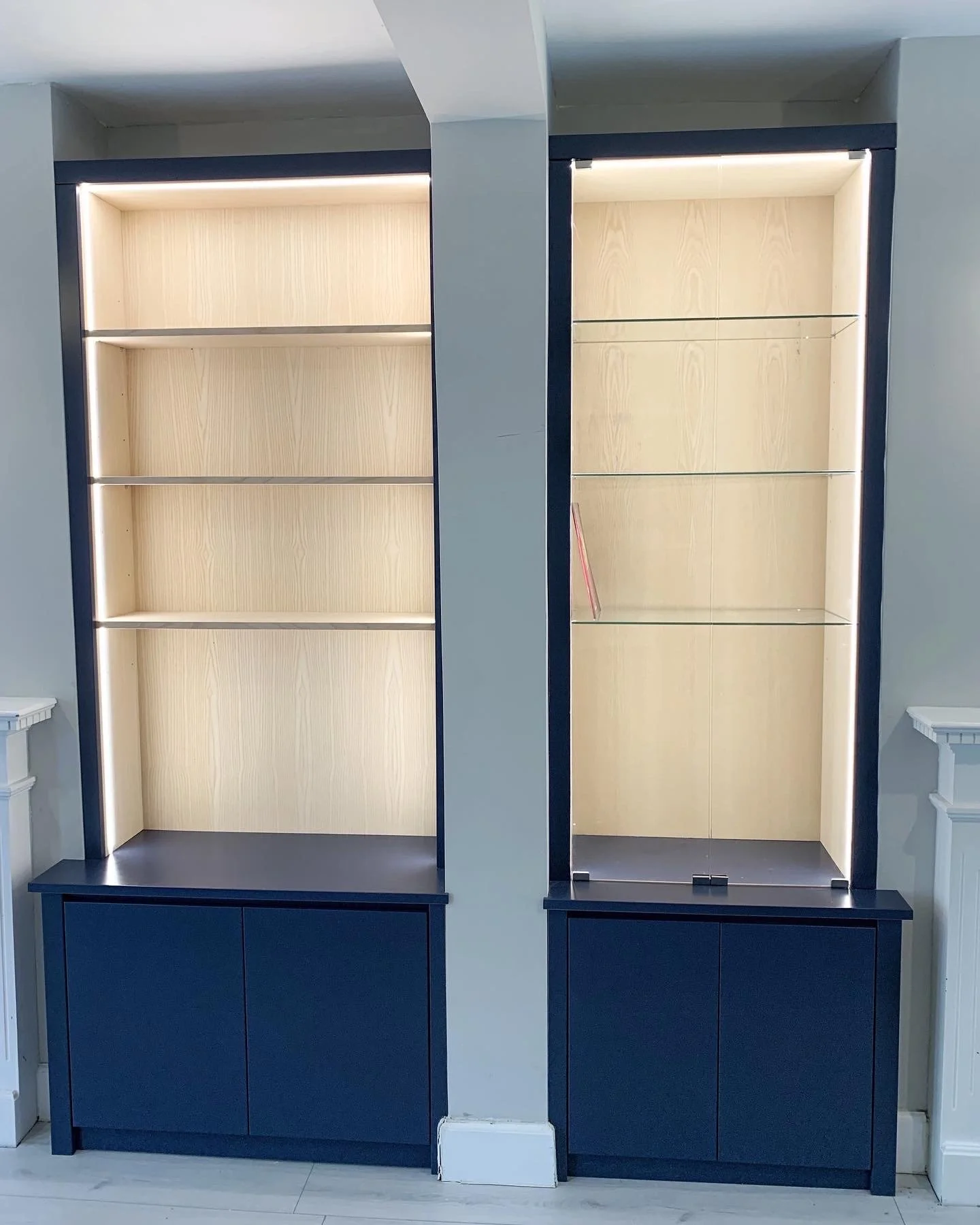 Two tall display cabinets with built-in lighting, empty shelves, and closed navy blue lower cabinets in a room with light-colored walls.