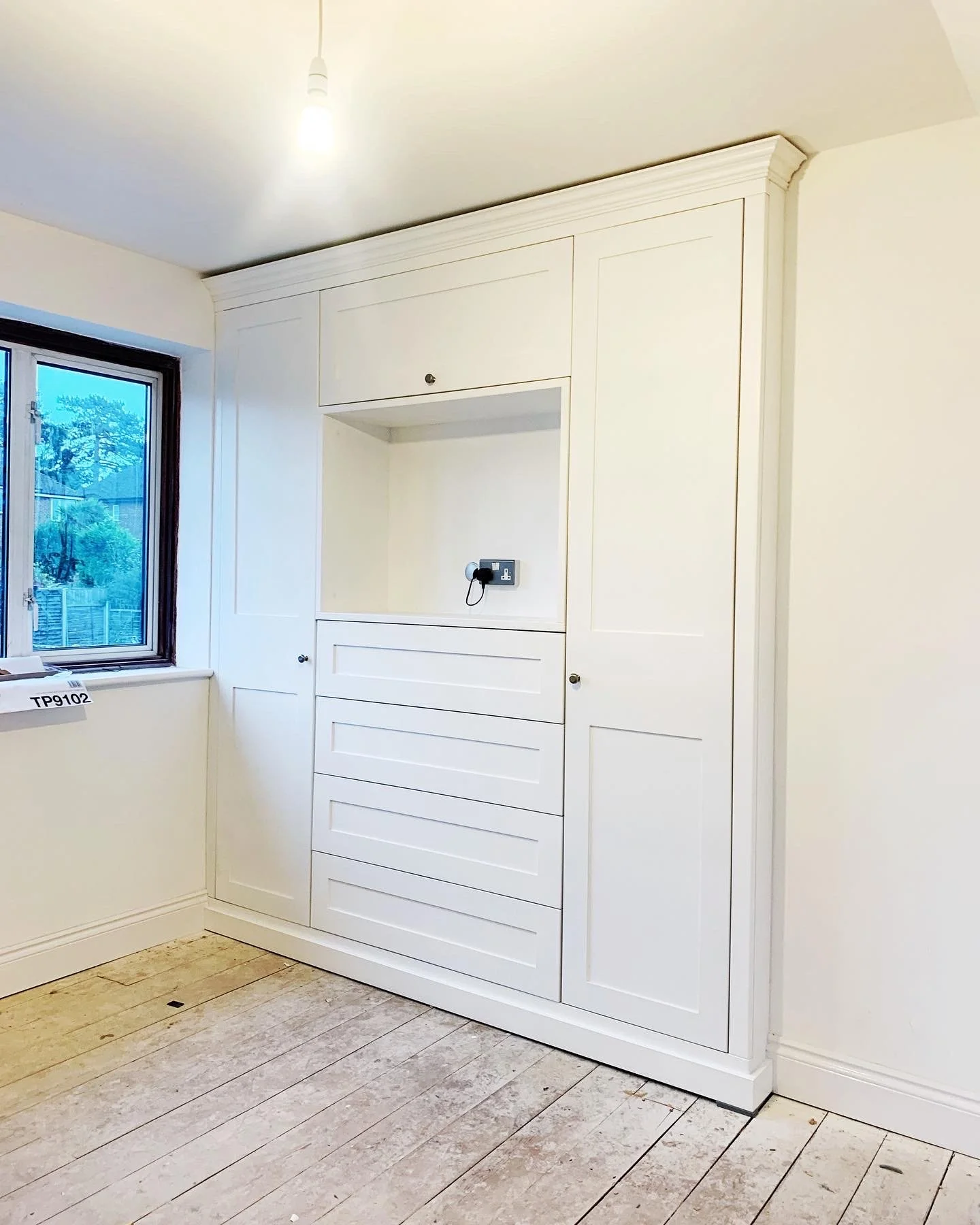 White built-in cabinet with drawers and doors, electrical outlet inside empty space, in a room with wooden floor and a window.