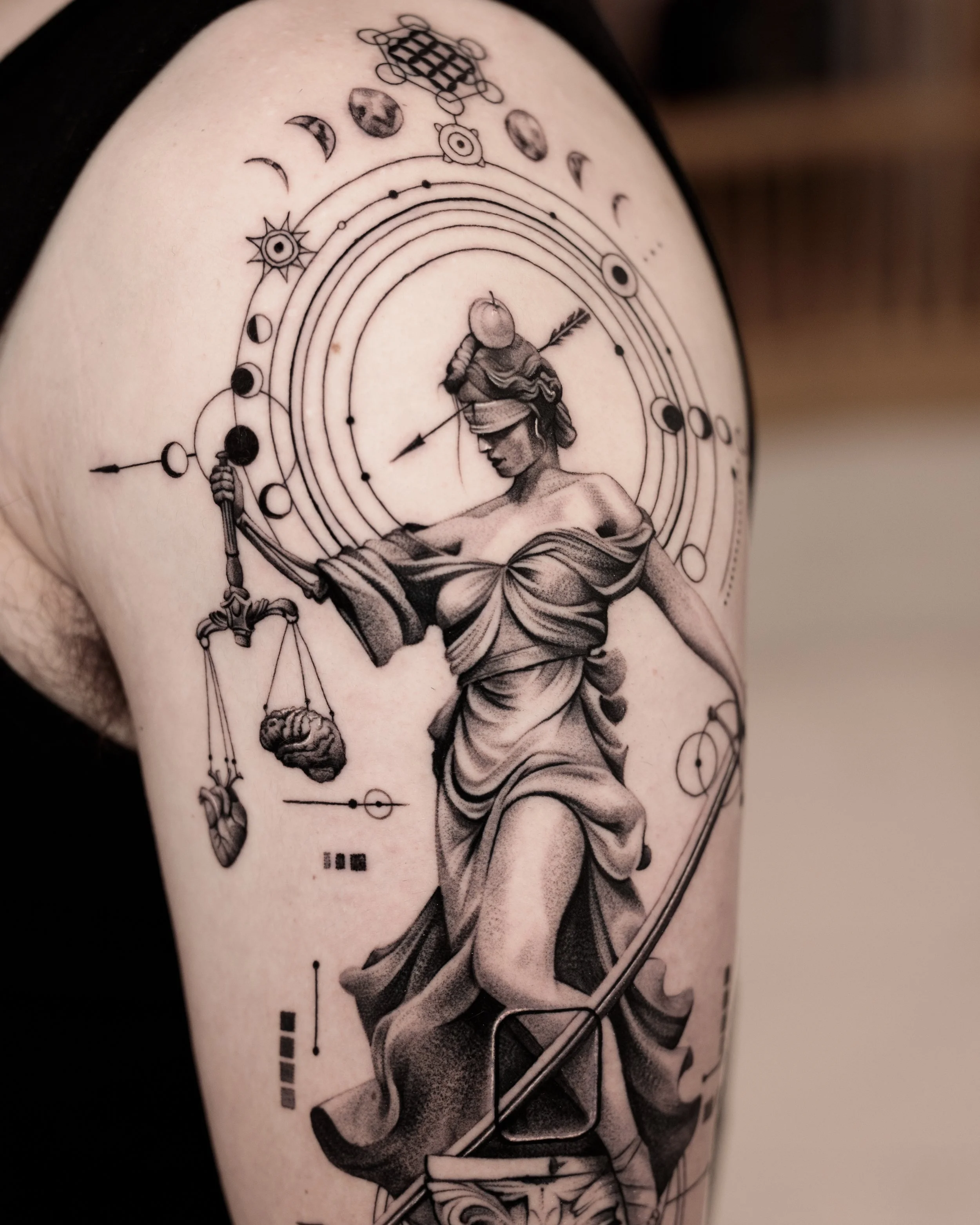 Black and grey upper arm tattoo of Lady Justice holding scales and a sword, surrounded by concentric circles, moon phases, and geometric details.
