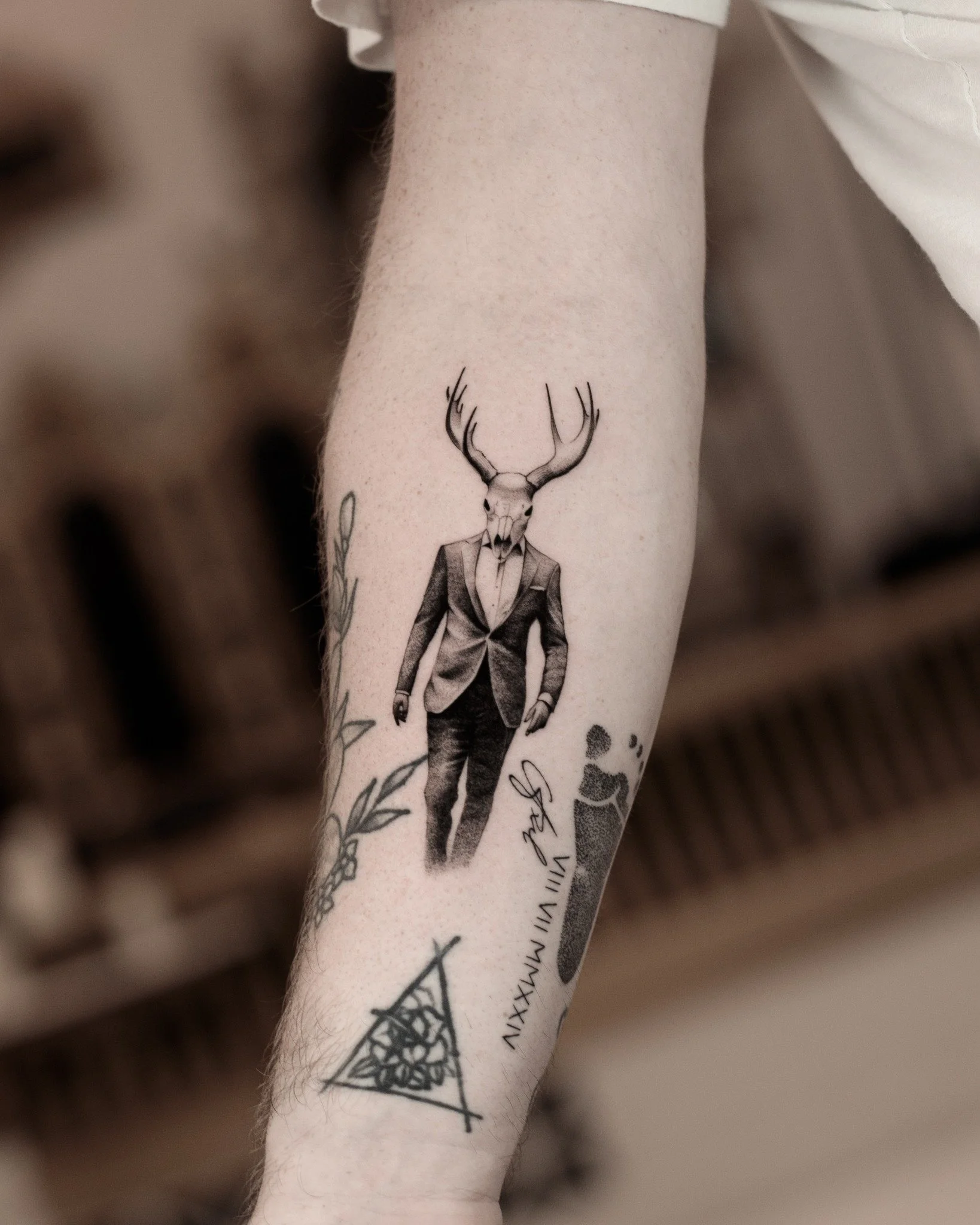 Black and grey microrealism tattoo of a figure wearing a suit, with an antlered deer skull in place of its head.