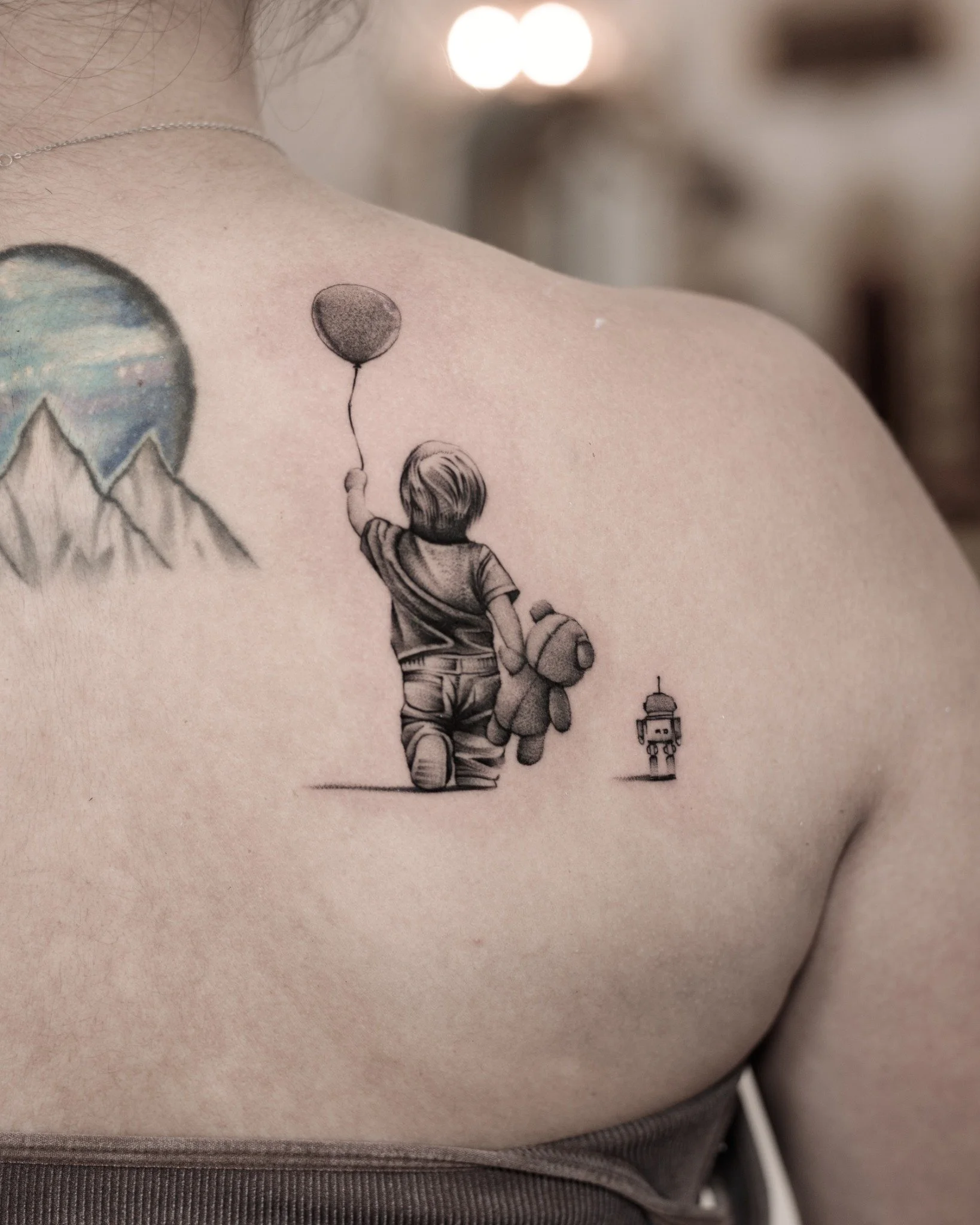 Black and grey upper back tattoo of a child seen from behind, holding a balloon and teddy bear, with a small toy robot beside them.