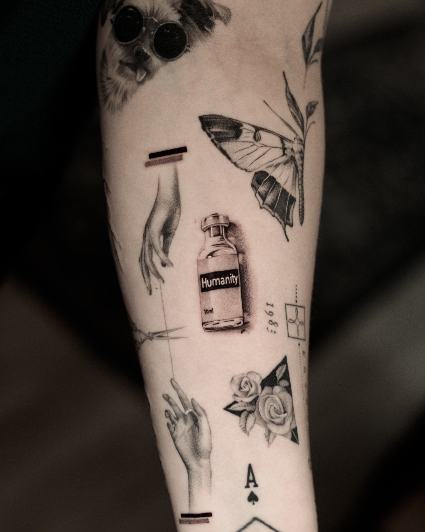 Black and grey microrealism tattoos of a glass vial labeled Humanity and two reaching hands, surrounded by small illustrative designs.