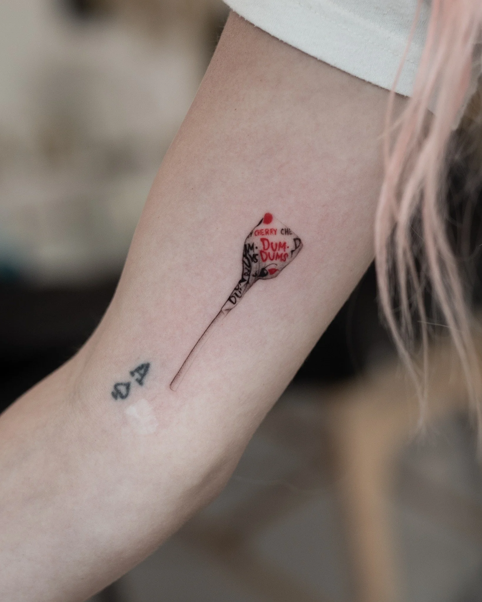 Realistic tattoo of a tiny wrapped cherry Dum Dums lollipop, with red and black lettering and a long, slender stick.