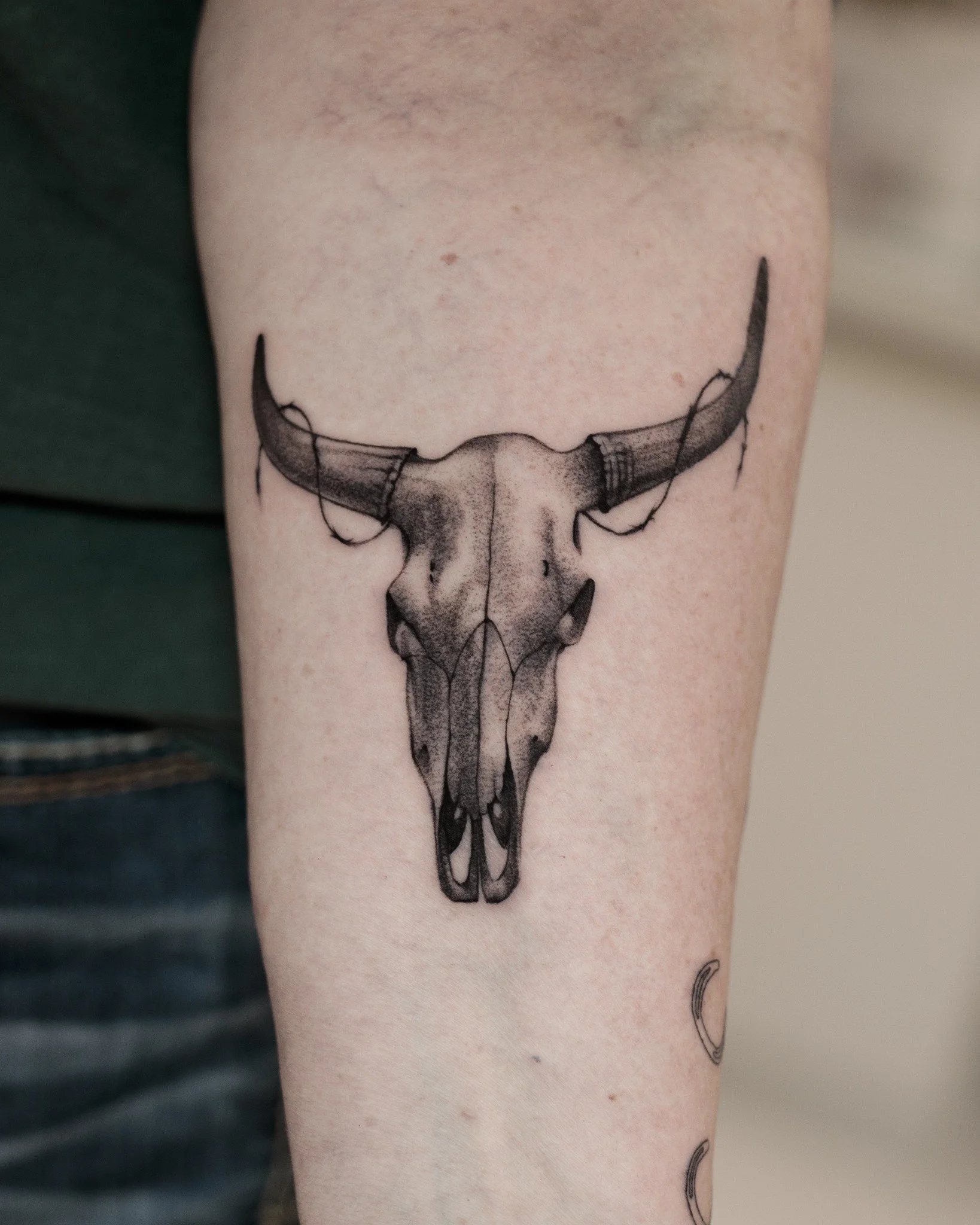 Black and grey tattoo of a bull skull with curved horns wrapped in loops of barbed wire.
