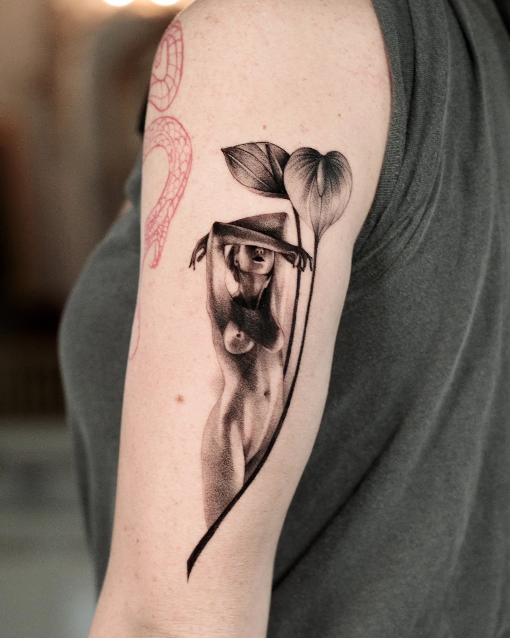 Black and grey upper arm microrealism tattoo of a woman in a wide-brimmed hat, framed by two leaves and a sweeping curved stem.